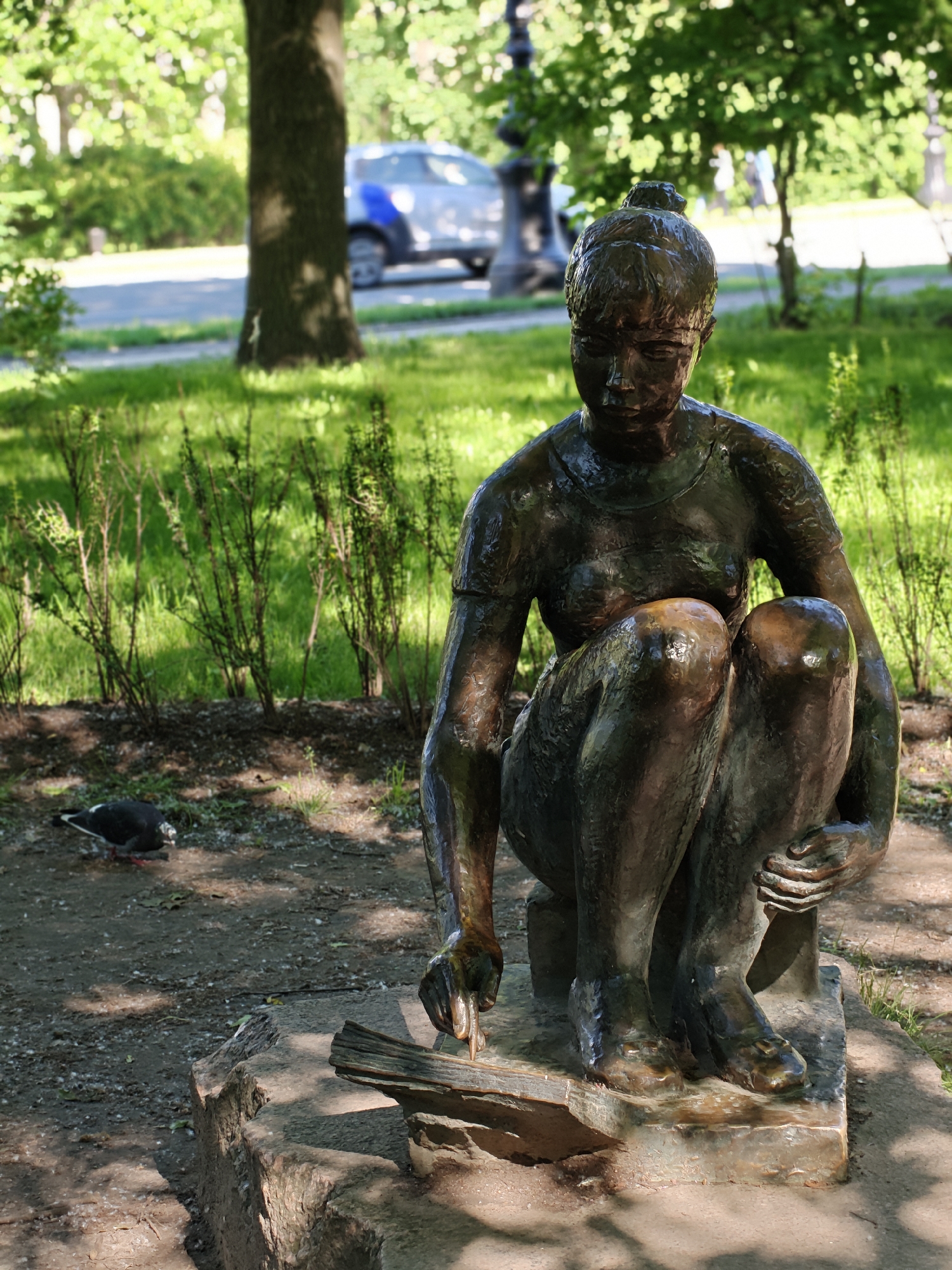 A bronze statue of a young woman, depicted in a crouching or kneeling posture, is situated outdoors in a park setting. The statue is dark bronze in color and appears to have a textured surface, reflecting ambient light. The woman's hair is pulled back into a bun. Her gaze is directed downward. Her arms are bent, and her hands are positioned in her lap or on her knees. She is wearing a simple, round-neck garment that resembles a t-shirt.
The statue rests on a rough-hewn stone base, which itself is placed on a patch of dirt and scattered leaves. To the left of the statue, a pigeon is foraging on the ground.
The background of the image features lush green grass and trees, suggesting a well-maintained park or garden. Sunlight filters through the leaves, creating dappled shade on the ground. In the middle ground, a light-colored vehicle with blue accents, possibly a car or van, is visible on a paved surface, suggesting a road or pathway is nearby. The overall impression is that of a public space in a city, likely Saint Petersburg, Russia, given the architectural style of lampposts visible in the background, which are characteristic of the city.