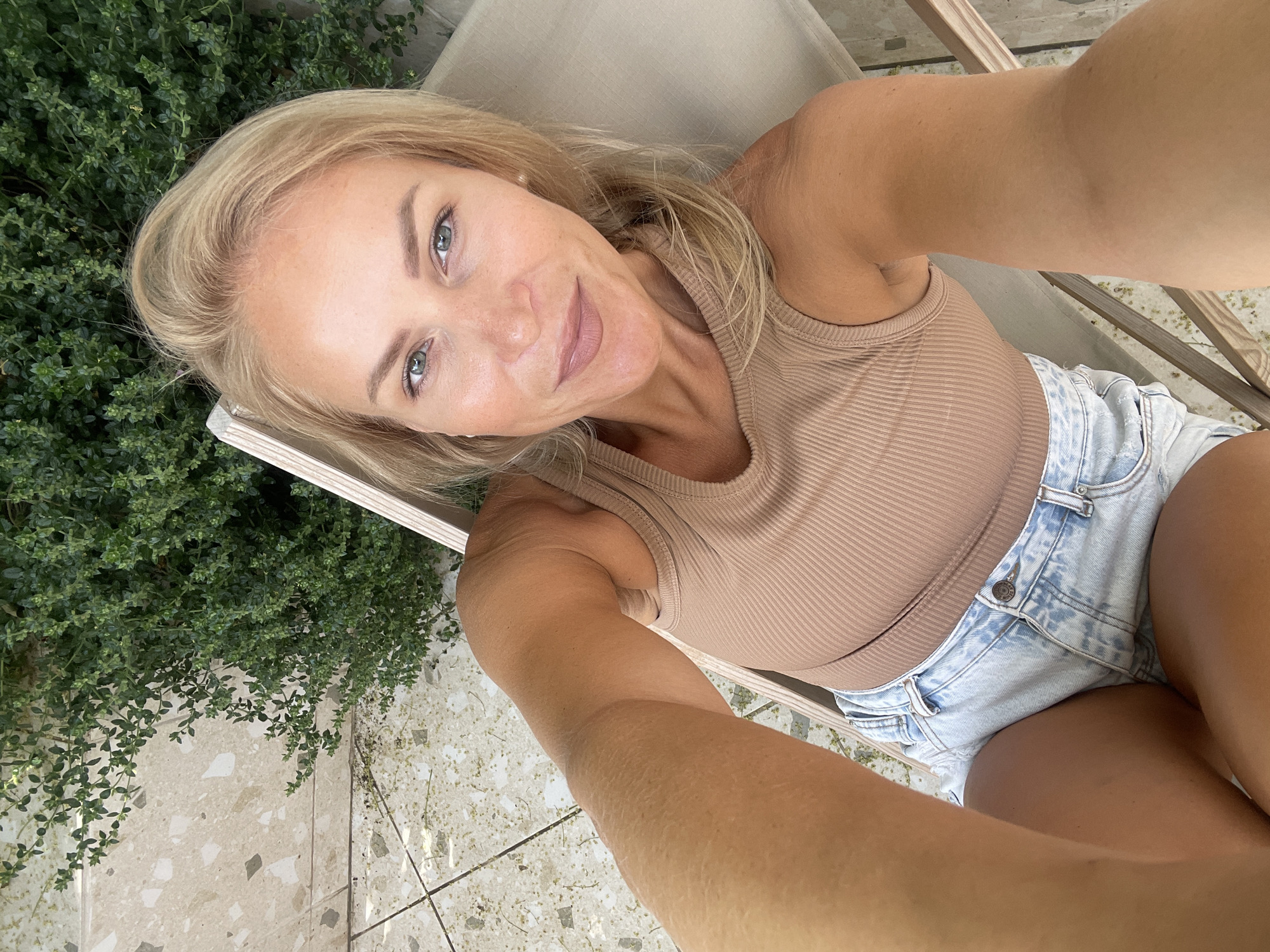 In this image, a blonde woman with light blue eyes is captured in a selfie. She is seated on a beige lounge chair outdoors, with a dense green bush behind her and a terrazzo-tiled floor in the foreground. The woman is wearing a light brown ribbed tank top and distressed denim shorts. She has light makeup on, including lipstick and mascara. Her right arm is extended, holding the camera, and her left arm is resting on her lap. The overall scene suggests a relaxed, sunny day.