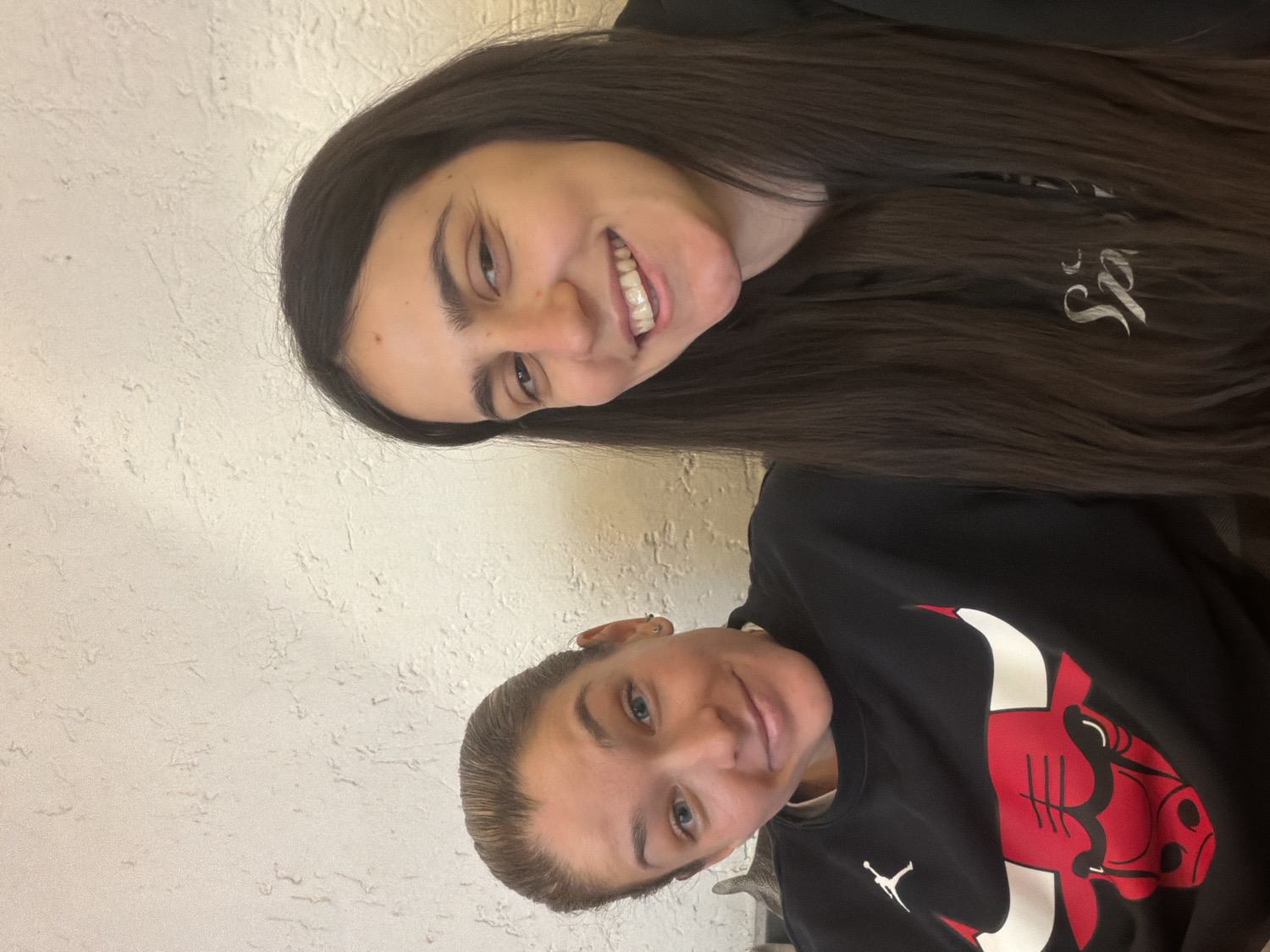 The image features two young women captured in a close-up, head-and-shoulder portrait, oriented on its side (rotated counter-clockwise). The woman in the upper portion of the frame has long, dark brown hair, styled straight, and is smiling broadly, revealing her teeth. She wears a dark long-sleeved top with a faint white cursive logo, possibly starting with 'J' or 'S', visible on her chest area. Below her, in the lower portion of the frame, is another woman with lighter, shorter brown hair, pulled back. She smiles subtly towards the camera and is wearing a black sweatshirt. The sweatshirt prominently displays the red and white Chicago Bulls logo on the front, along with a smaller white Jumpman (Jordan brand) logo below it. Both subjects appear happy and friendly. The background is a textured, light-colored wall, possibly stucco or plaster. The setting is indoors, with general ambient lighting. No specific time of day or weather conditions can be determined from the image. The location context provided is Sofia, Bulgaria, although no specific visual cues within the image confirm this.
