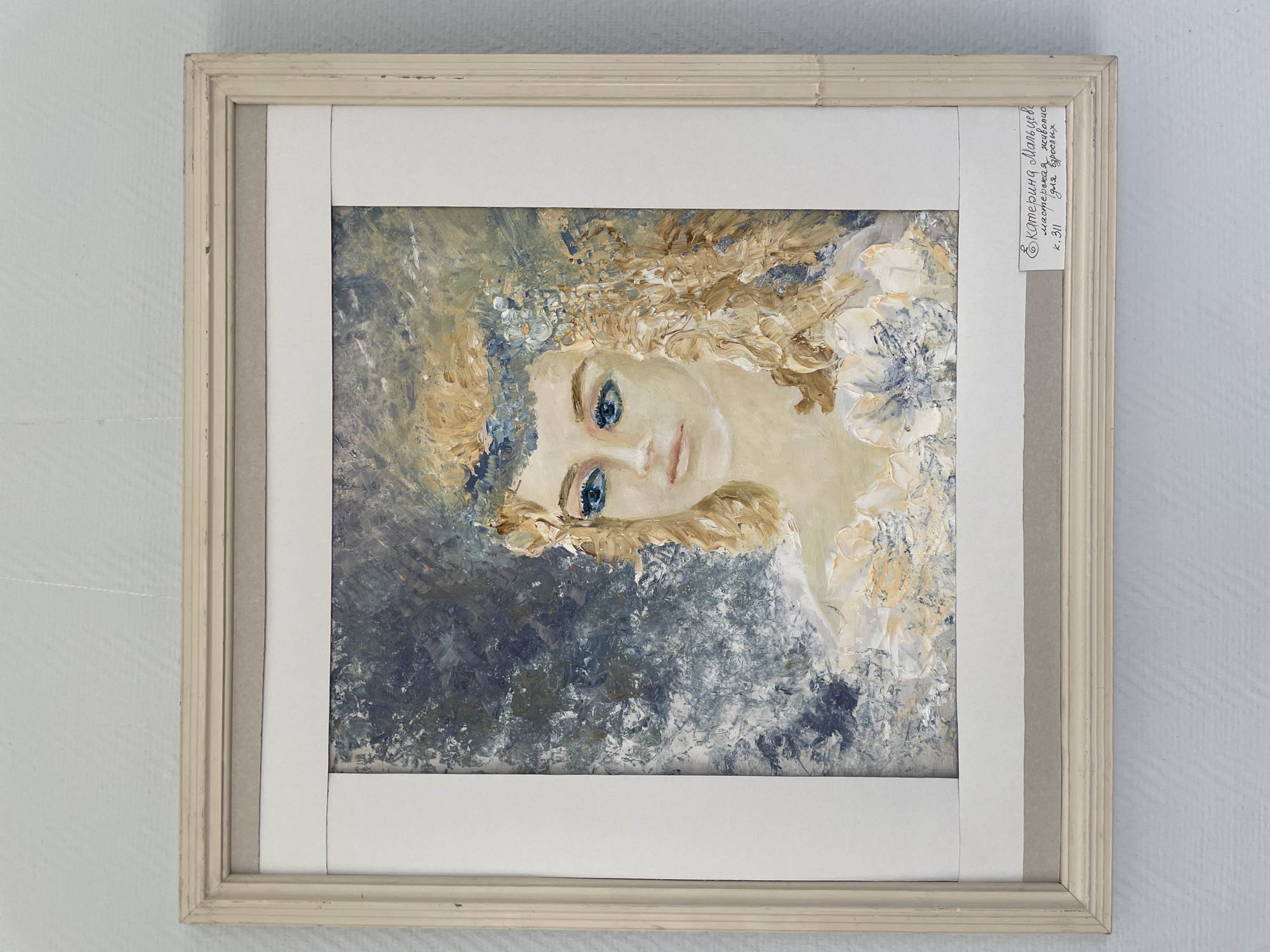 A framed painting of a woman with blonde hair and blue eyes is displayed against a light blue, textured wall. The painting is in a light wood frame with a cream-colored mat. A small, handwritten label in Russian is affixed to the bottom right of the mat. The label reads: "Е. Катерина Мальцева мастерская живопись к. 311 для взрослых". Based on the inscription, this painting is likely located in a workshop or studio related to fine arts in Perm, Russia.