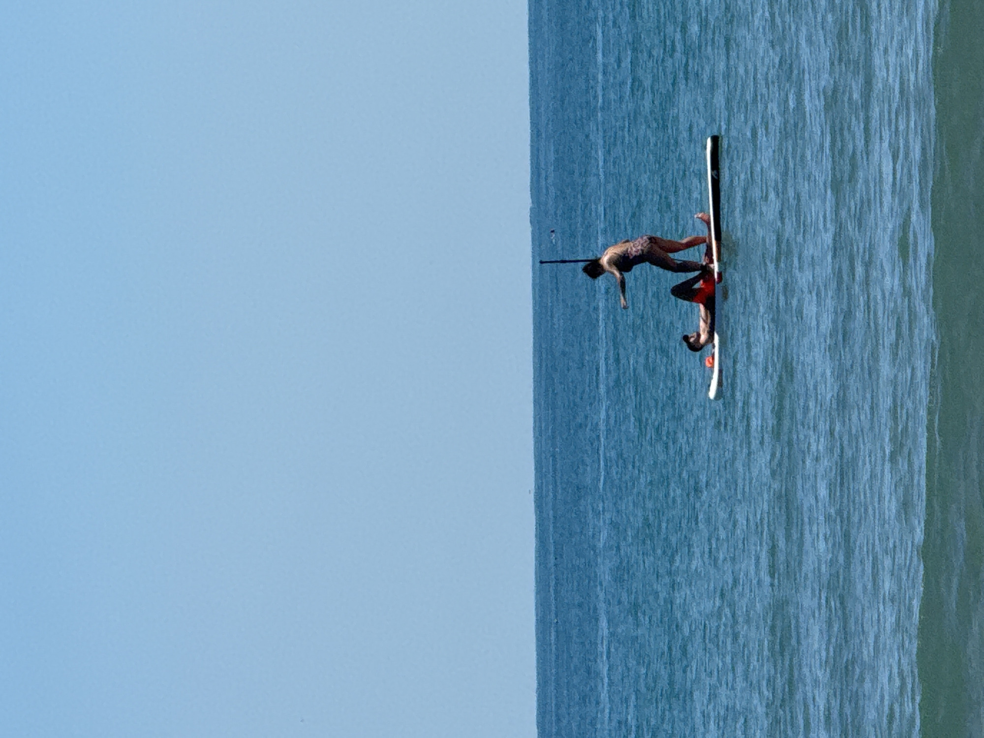 Two individuals are on a paddleboard in the sea off the coast of Sozopol, Bulgaria. One person is standing and appears to be in motion, with their left arm extended. The other person is lying down on the paddleboard, facing upwards. The water is a deep blue, and the sky is a clear, light blue. There are small waves on the surface of the water, and the horizon is visible in the distance.