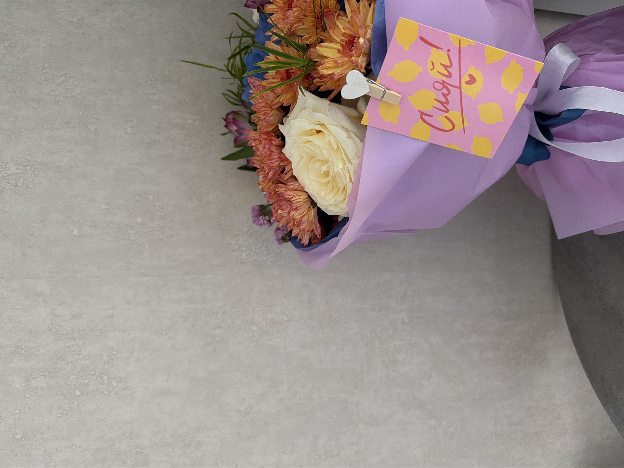 A bouquet of flowers, wrapped in light purple paper, is displayed against a textured, light gray wall. The bouquet contains orange chrysanthemums, a cream-colored rose, and some greenery. A small, pink card with yellow lemon illustrations and red lettering is attached to the bouquet with a wooden clothespin. The lettering on the card is in Russian and reads "Сиди!" which translates to "Sit!". The background wall has a slightly mottled appearance, suggestive of concrete or stucco. The bottom of the image shows a dark gray surface, possibly a table or chair. There are no people or animals present in the image. The scene is indoors, likely in a residential or commercial setting. The location of Petropavlovsk-Kamchatsky, Russia is not determinable from the visual information provided in the image.