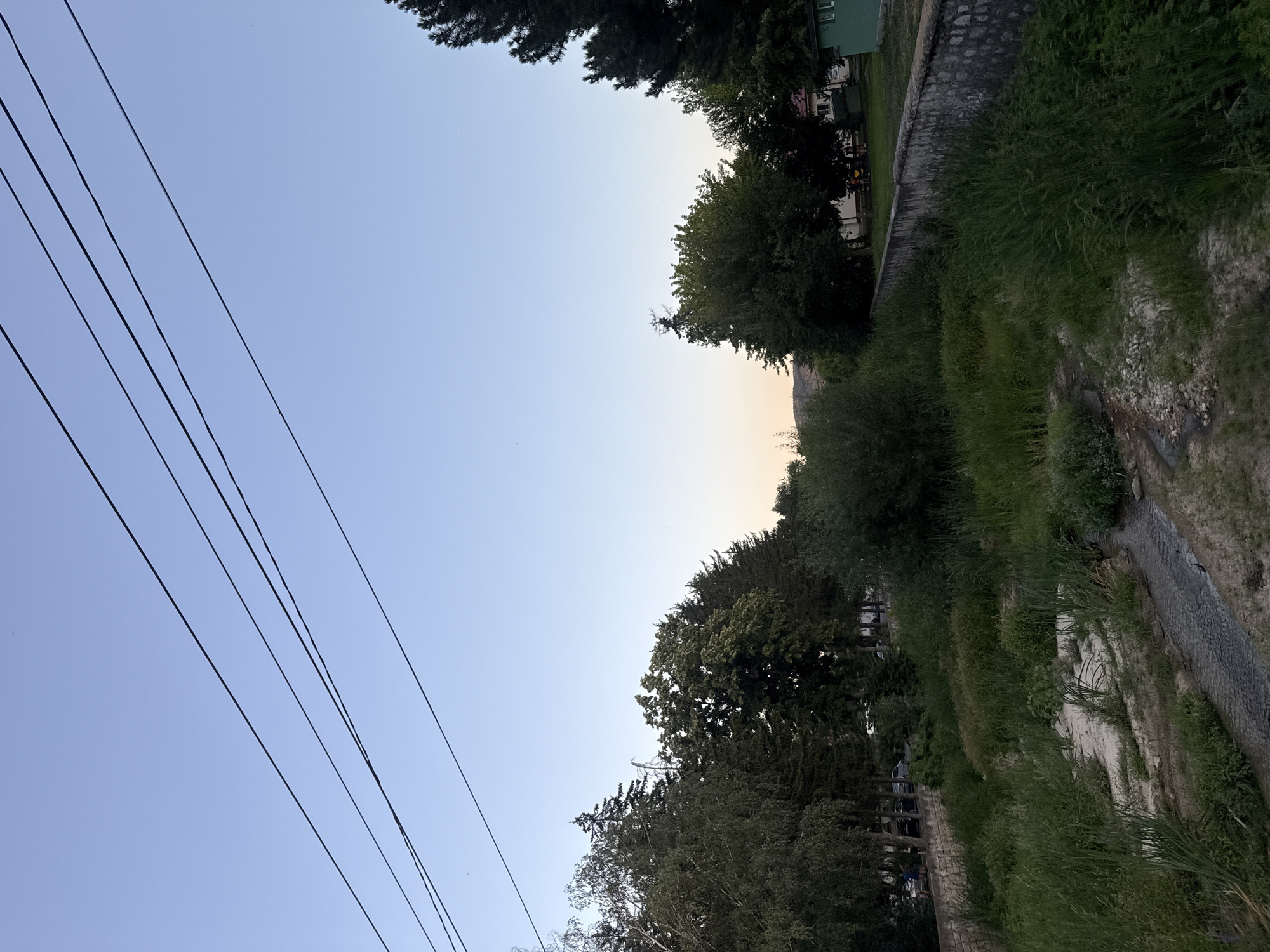 An overhead shot captures a serene riverbed in Strelcha, Bulgaria, under a clear, pale blue sky. Several electrical wires crisscross the upper portion of the frame, contrasting with the natural landscape below. The riverbed is a mosaic of dry sand and pebbles, edged by lush green grass and reeds. A narrow stream of water trickles through the center. The banks of the river are lined with a stone retaining wall. Beyond the wall, a dense line of trees, varying in height and foliage, creates a verdant backdrop. In the distance, peeking through the trees, are hints of buildings and a small green structure. The scene evokes a sense of quietude and natural beauty within an urban setting.