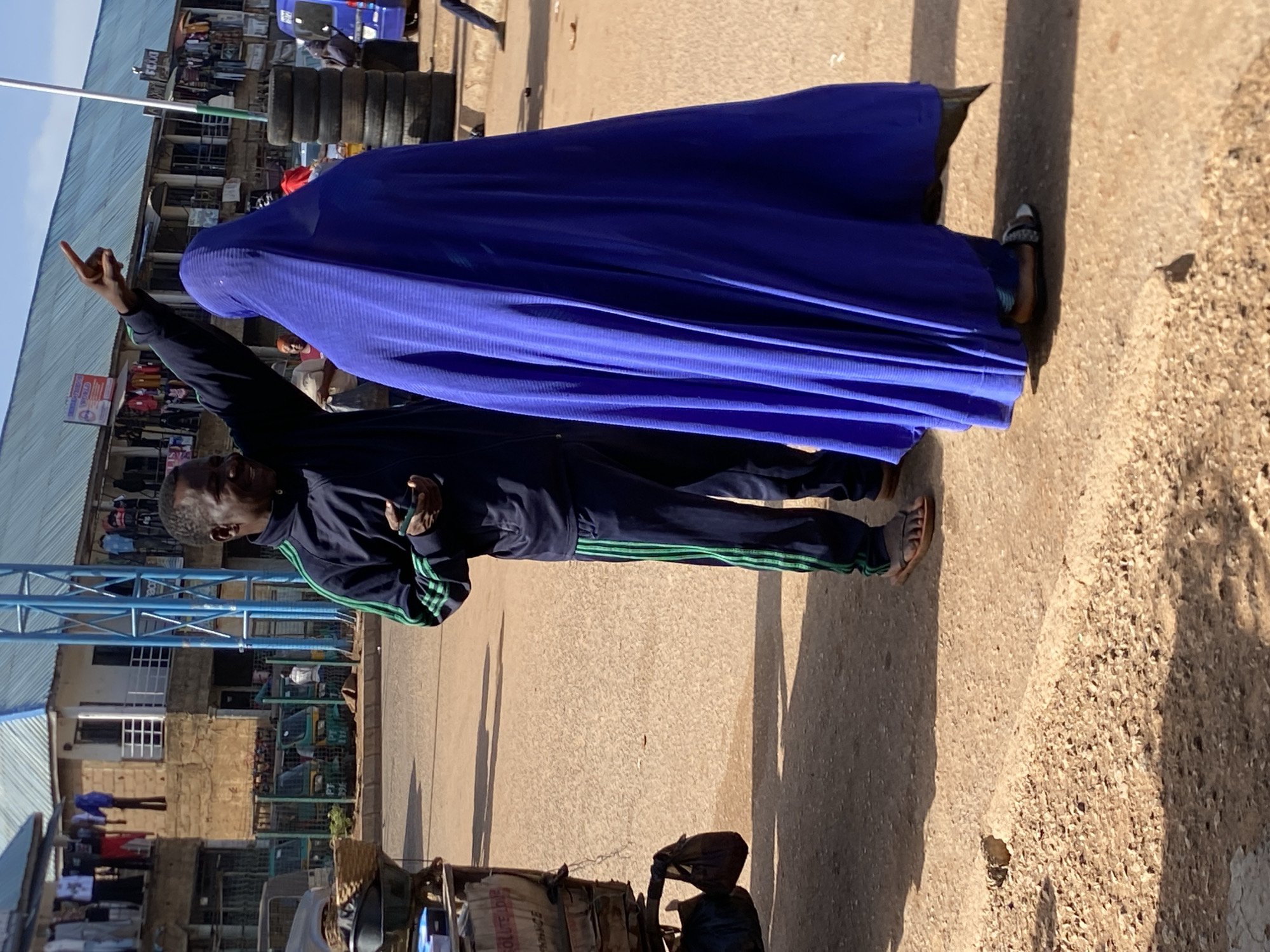 The image, oriented vertically due to a 90-degree clockwise rotation, depicts an outdoor market or street scene in Bukuru, Nigeria, under bright daylight. Long shadows cast on the ground suggest it is either morning or late afternoon.
Two individuals are the main subjects in the foreground. On the left, a person wearing a dark blue tracksuit with distinctive green stripes along the sleeves and pant legs stands, looking towards the right. Their right hand is raised, making a gesture with the index and pinky fingers extended (often interpreted as a "rock on" or similar sign), while they hold a mobile phone in their left hand. To their right, and slightly behind, stands another person fully enveloped in a long, flowing royal blue garment that covers their entire body and head. Only their feet, wearing light-colored sandals or slippers, are visible beneath the garment.
The background reveals a bustling market environment. Various buildings with corrugated metal roofs and walls, some appearing to be brick or concrete, line the street. A large stack of black vehicle tires is visible on the right side of the background. Further back, several light-colored tricycle taxis, commonly known as "keke napeps" in Nigeria, are parked. A blue metal frame structure, possibly part of a stall or signage, is also present. The ground is rough, appearing to be a mix of dirt and gravel. In the lower left foreground, a cart or stand laden with goods, including a white sack with some illegible text, is partially visible. A white sign in the mid-background has the word "FLEA" partially discernible on it.