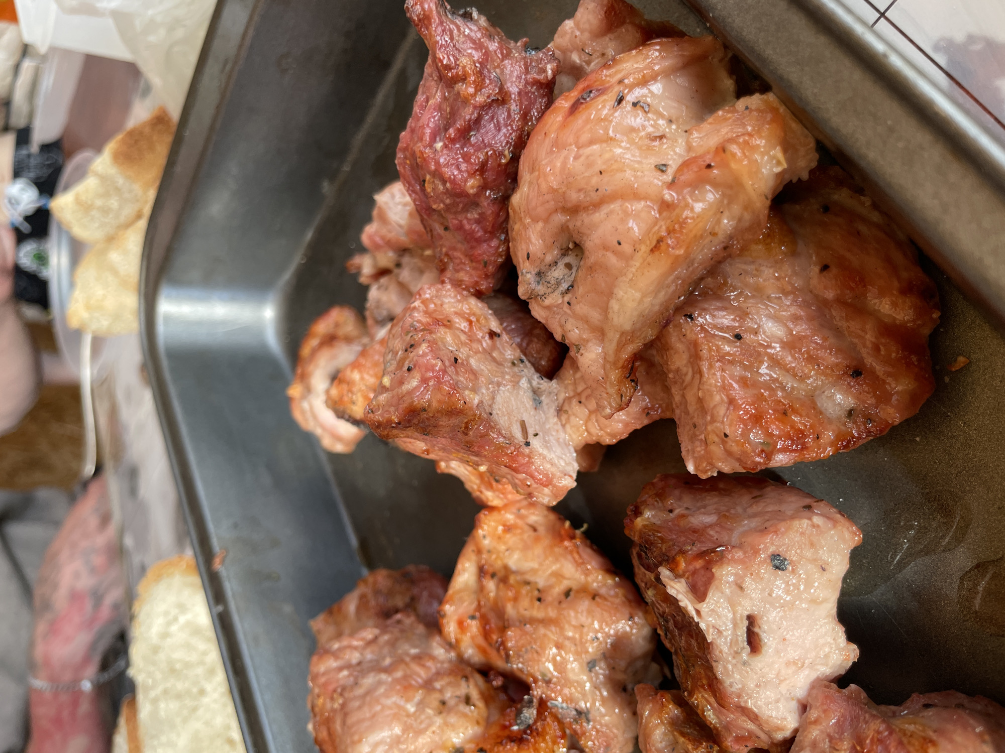 A close-up shot shows a metal pan filled with cooked pieces of meat, likely pork, seasoned with herbs and spices. The meat appears to be in bite-sized cubes and has a glistening, slightly browned exterior, indicating it has been roasted or pan-fried. In the background, out of focus, there are slices of bread and the hands of two people, one with tattoos on their arm. The overall setting suggests a meal being prepared or served, possibly at an outdoor gathering or casual dining environment. The image lacks any specific visual cues that would associate it with Novokuznetsk, Russia, or any particular city.