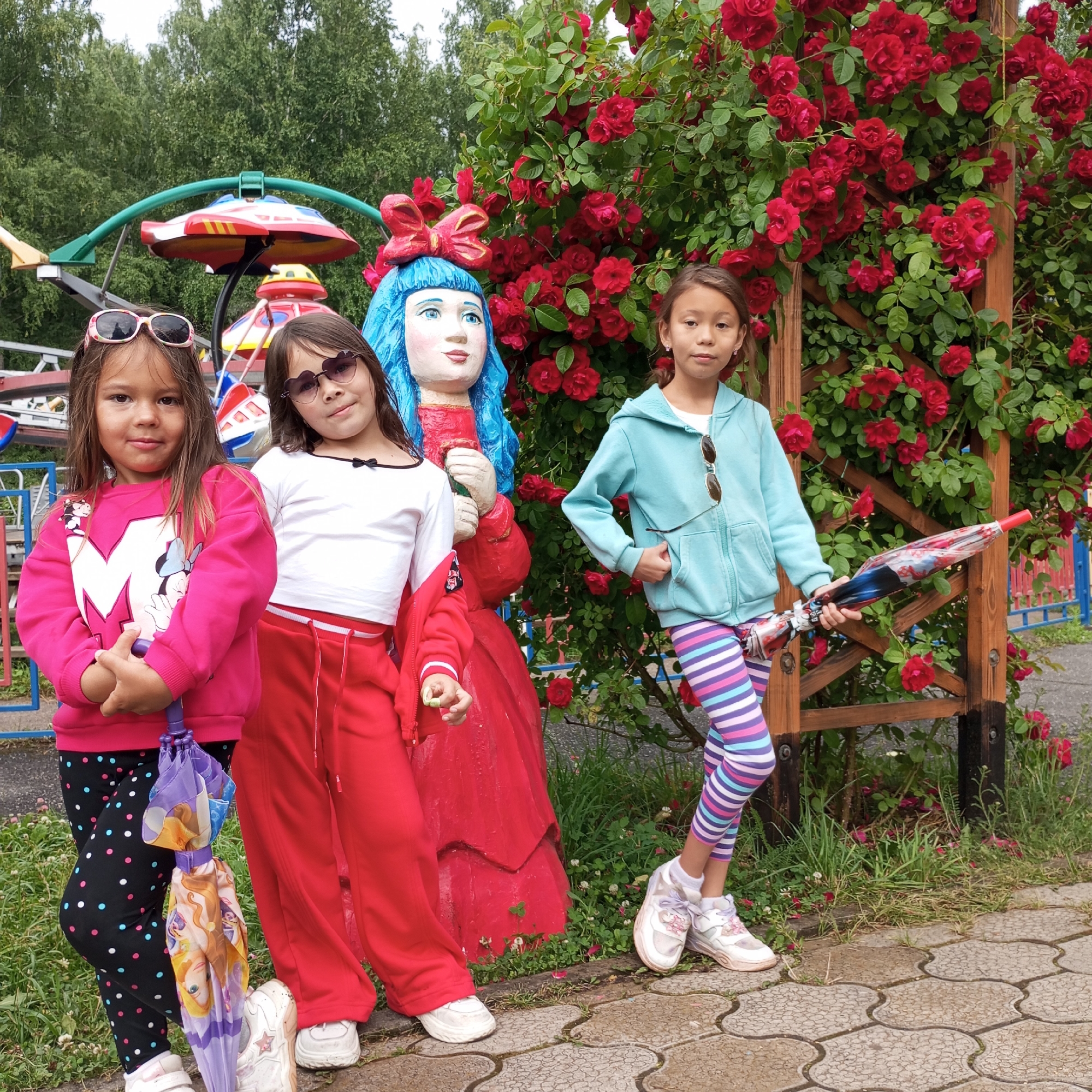 Three young girls are posing for a photo in an outdoor park setting, likely during the daytime given the natural light. Behind them is a large bush covered in vibrant red roses and a wooden trellis. To the left, parts of a colorful amusement park ride are visible, suggesting the location is an amusement park or a recreational area.
The girl on the left is wearing a bright pink sweatshirt with a white "M" and a Minnie Mouse graphic, black leggings with colorful polka dots, and pink and white sneakers. She is holding a closed purple umbrella decorated with images of princesses. She has dark hair and is wearing sunglasses.
The middle girl is wearing a white long-sleeved shirt with a black bow detail at the neckline, red tracksuit pants, and white sneakers. She has dark hair and is wearing heart-shaped sunglasses.
The girl on the right is wearing a light blue hooded sweatshirt, colorful striped leggings (pink, blue, and purple), and white sneakers. She has dark hair and is wearing sunglasses pushed up on her head. She is holding a closed umbrella with a pattern that includes red, white, and blue elements.
A large, somewhat weathered statue of a doll-like figure with blue hair and a red dress stands between the first two girls. The overall atmosphere appears casual and cheerful, with the girls looking directly at the camera, some with smiles. The ground is paved with interlocking decorative stones, some of which appear wet, hinting at recent rain or a damp surface. The presence of a park ride and lush greenery suggests a pleasant, possibly warm, day for outdoor activities.