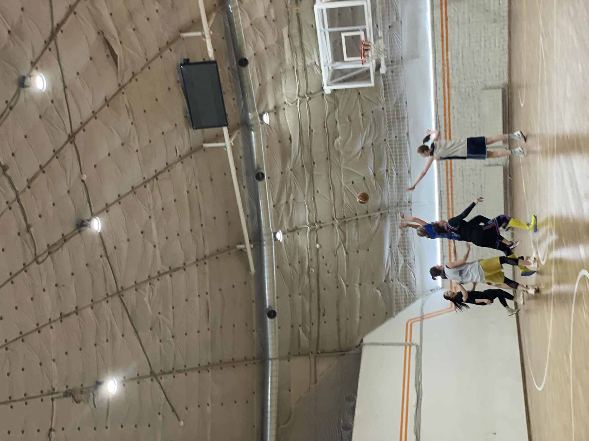 The image shows an indoor basketball court with a high, rounded ceiling covered in padded material. A basketball game appears to be in progress, with multiple players visible on the court. One player is reaching for a basketball in mid-air. A basketball hoop and a scoreboard are mounted on the wall. The location is a basketball court in Saint Petersburg, Russia.