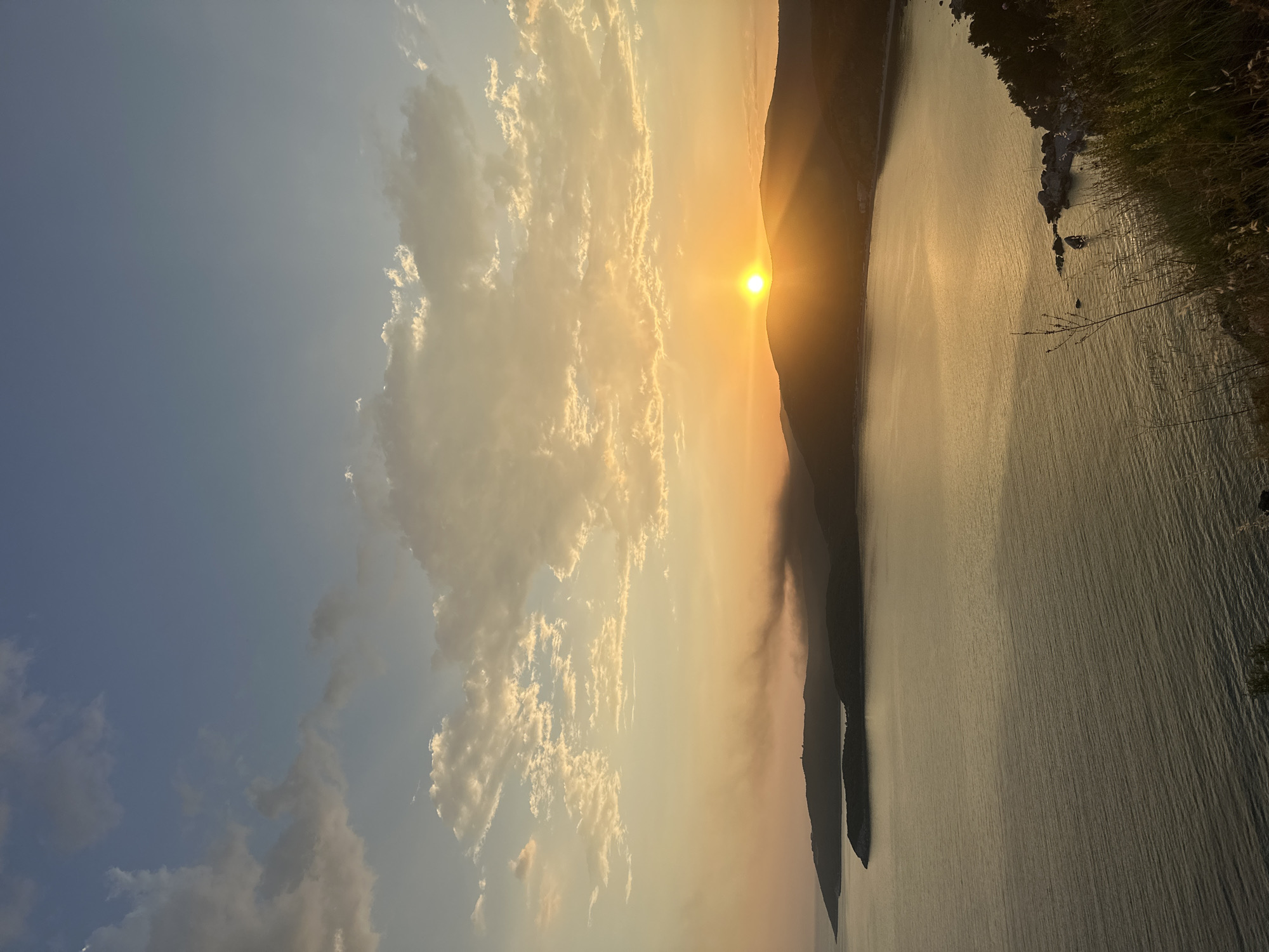 The image depicts a sunset over the Adriatic Sea, likely near Budva, Montenegro. The sun is positioned behind a coastal mountain range, casting a warm, golden glow across the sky and water. Clouds are scattered across the sky, illuminated by the sunset. The sea surface reflects the sunlight, creating a shimmering effect. Vegetation is visible on the shoreline.