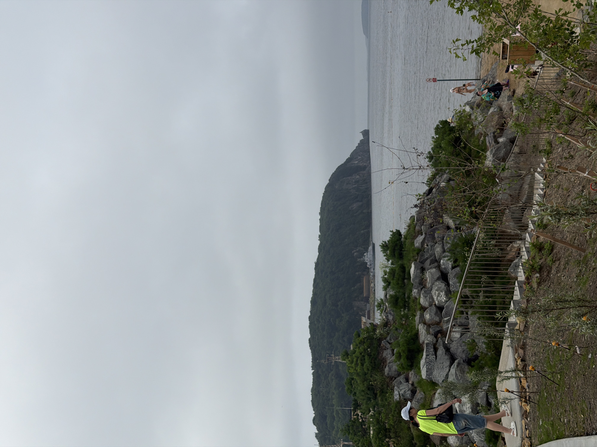 This is a photograph taken on a cloudy day at a coastal park area in Vladivostok, Russia, likely on Russky Island given the presence of a naval ship and the rugged coastline. In the foreground, a person wearing a bright yellow-green t-shirt, denim shorts, a white cap, and white sandals walks away from the camera on a paved path. To the right, a metal handrail leads up a set of stairs, and beyond that, the coastline features rocky formations interspersed with green shrubs and trees.
In the mid-ground, the sea stretches out, meeting a lush, green, tree-covered hill that slopes down to the water. A naval vessel is visible in the distance near the base of the hill. Several individuals are present on a sandy beach area further to the right, including a woman in a bikini, another woman in a green and black garment sitting on rocks, and a child. A red and white warning sign stands on a pole near the water's edge. The overall environment is natural, with a mix of rocky terrain, vegetation, and the sea under an overcast sky.
