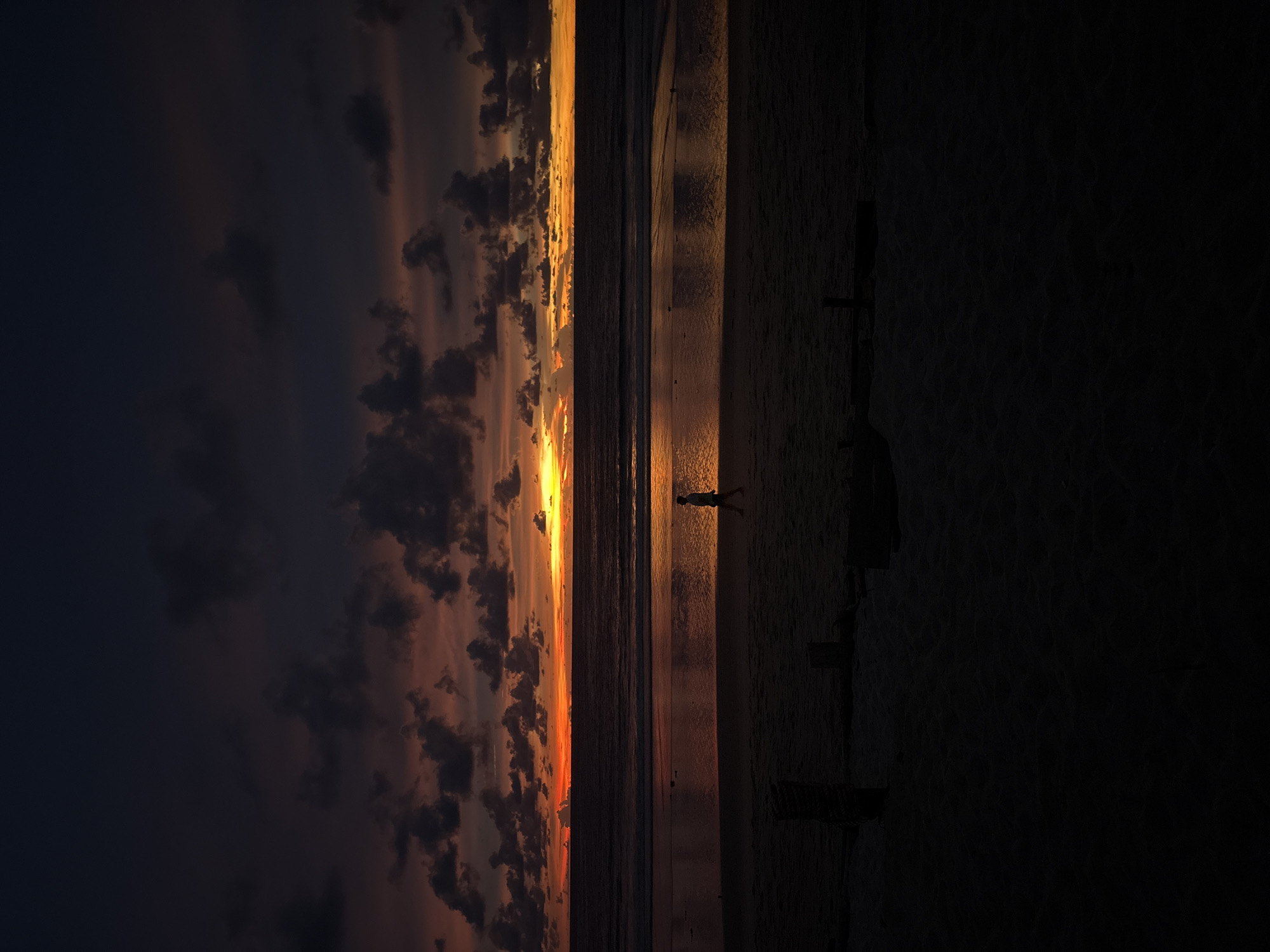 The image depicts a sunset scene at a beach, likely in Choeng Thale, Thailand, given the dark sand and typical coastal landscape. A lone figure, appearing to be a child, walks along the wet sand at the edge of the ocean. The sky is filled with dramatic, dark clouds interspersed with vibrant streaks of orange and yellow from the setting sun. The ocean reflects the colors of the sunset, creating a shimmering effect. In the foreground, the dark sand is textured with footprints and appears to have a few wooden posts or remnants of structures. The overall atmosphere is serene and slightly somber due to the fading light and deep shadows.