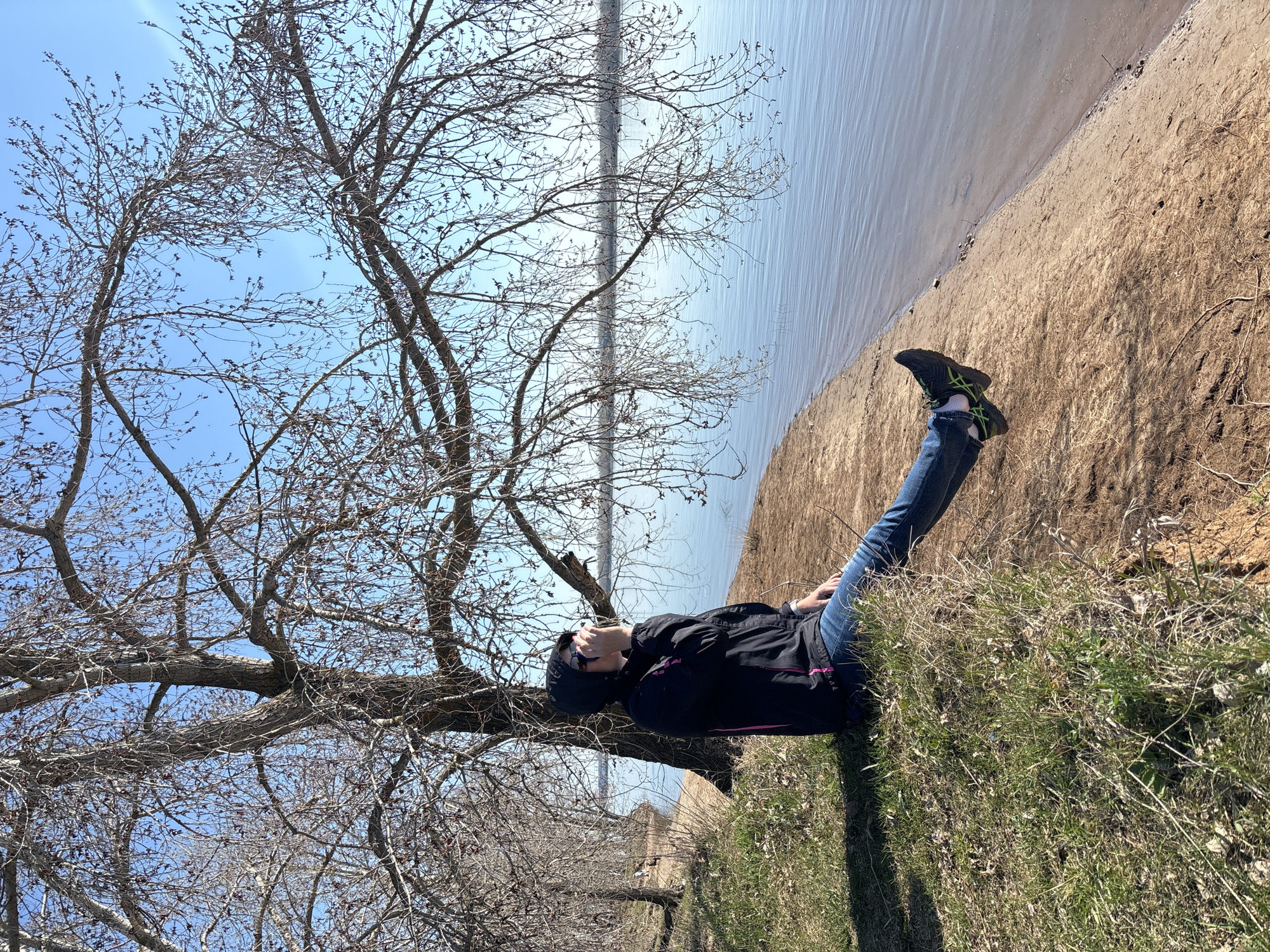 A person is lying on the grass near a body of water in Chaikovsky, Russia. The person is wearing jeans, black shoes with green accents, and a black jacket with pink trim. A bare tree is to the left. The body of water is to the right, with a sandy bank between the grass and the water. The sky is blue.