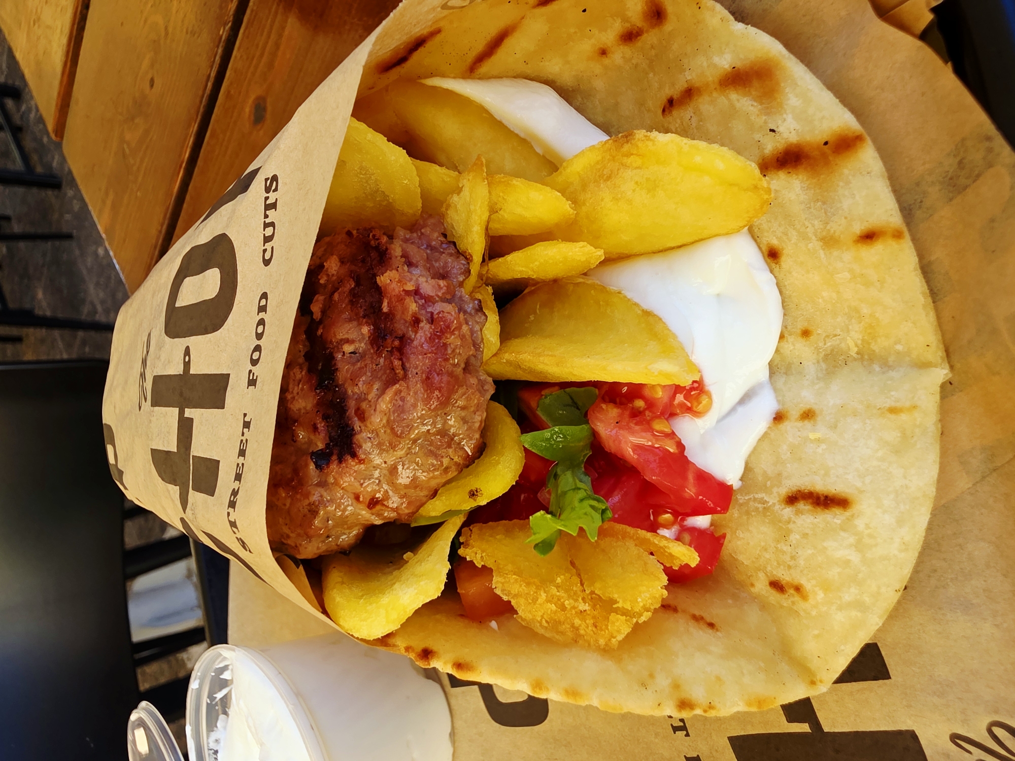 A close-up, overhead shot shows a large gyro wrapped in brown paper packaging printed with "HO STREET FOOD CUTS." The gyro is filled with a thick, seasoned meat patty, thick-cut fried potatoes, chopped tomatoes, lettuce, and a white sauce, possibly tzatziki. The gyro is resting on a piece of brown paper that is also printed with black text, part of which reads "HO." A small white plastic container with a clear lid, also filled with white sauce, is visible to the left of the gyro. The gyro and container sit on a wooden table, suggesting an outdoor dining setting. The background is blurred, but hints of dark chairs and more wooden tables indicate a restaurant or cafe environment, likely in Chania, Greece, given the context of the prompt. The lighting is natural, and the focus is sharp on the gyro, highlighting its textures and ingredients.