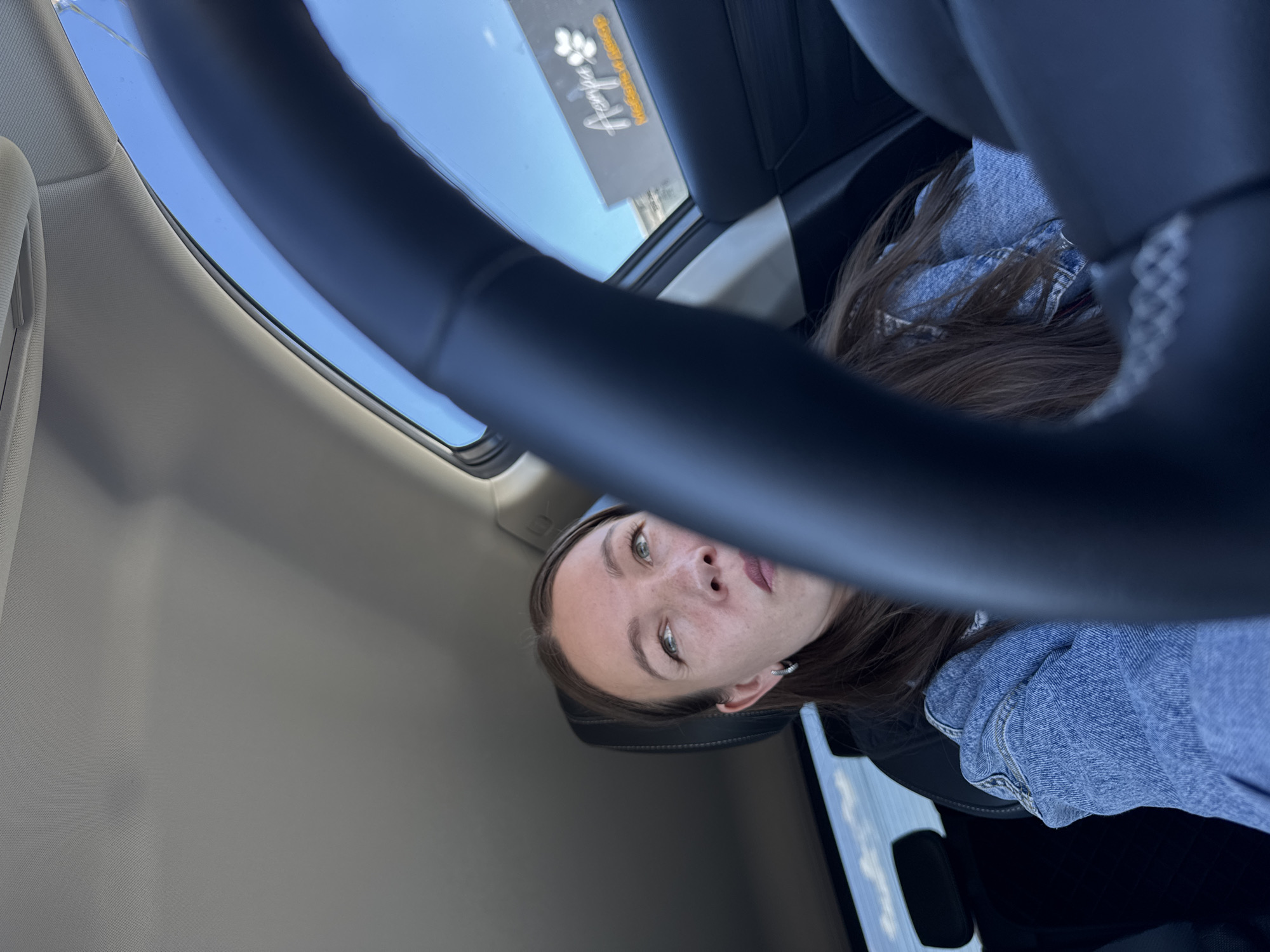 A young woman with fair skin, brown hair, and green eyes is seen from a low angle inside a vehicle. She is wearing a blue denim jacket. Above her, through an open sunroof, the blue sky is visible with a business sign that reads "Angela" in cursive script with paw print icons. The scene likely takes place in Chaikovsky, Russia, given the prompt. A black steering wheel is partially visible in the foreground. The interior of the vehicle is light grey.