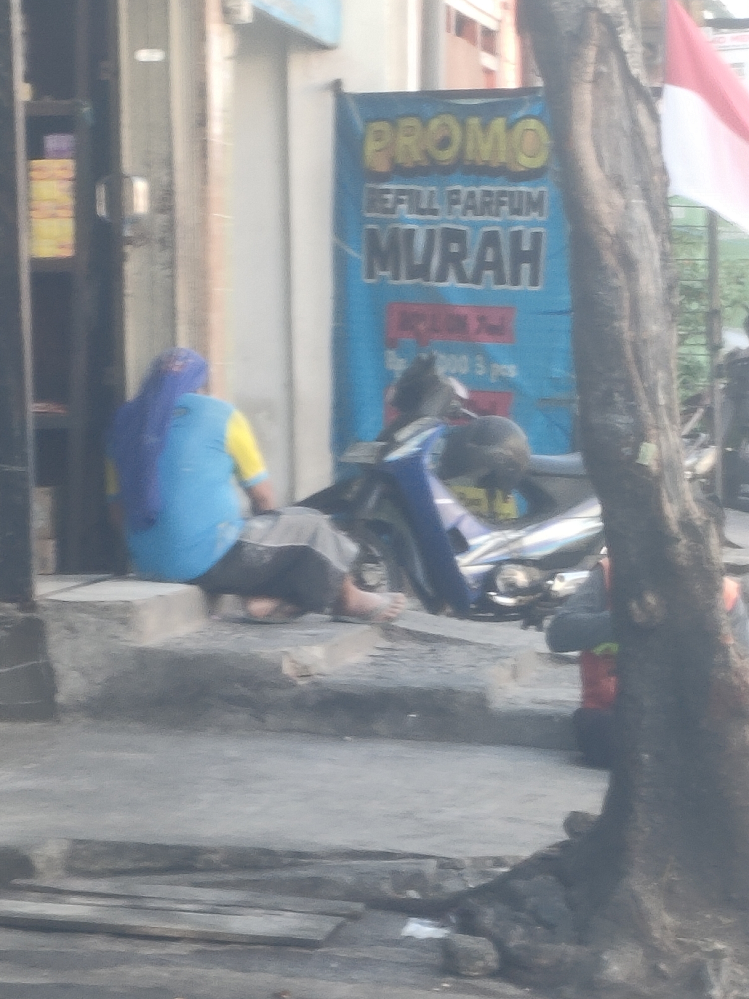 The image captures a street scene in Surabaya, Indonesia, likely in an urban commercial area. A person, possibly female, is seated on a raised concrete step in front of a building. They are wearing a blue headscarf and a two-toned shirt, half blue and half yellow. The person is facing away from the camera, and their lower body is obscured by the seat.
Behind the seated person, a motorcycle is parked. The motorcycle is predominantly blue and silver. To the right of the motorcycle, a large blue banner is visible, featuring yellow text that reads "PROMO" and smaller text below it. Further to the right, the trunk of a large tree with rough bark is prominently displayed, partially obscuring the background. The ground in the foreground appears to be asphalt or concrete, with some discarded debris. The overall image has a soft focus, suggesting it may have been taken with a low-resolution camera or from a distance. The lighting suggests an outdoor, daytime setting.