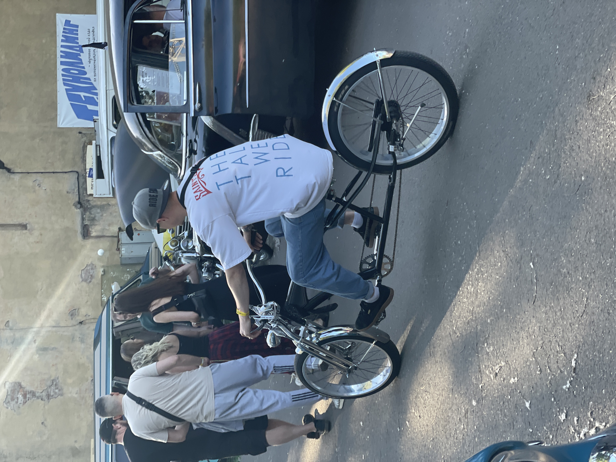 The image shows a man riding a low-slung custom bicycle. The man is wearing a white t-shirt with blue text that says "SAINT THE TAL WE RIDE", a grey baseball cap, and blue jeans. He is on a street with several vintage cars in the background. A banner with Russian text is visible on a building in the background, which reads "ТЕХНОАЛМАЛИНГ". The street appears to be paved with asphalt, and there are shadows cast by the sun. The overall setting suggests an outdoor event or gathering, possibly a car show, in Saint Petersburg, Russia, given the Russian text on the banner. There are other people visible in the background, some standing and looking at the cars or interacting with each other. The atmosphere seems casual and relaxed, with a focus on retro vehicles and custom creations.