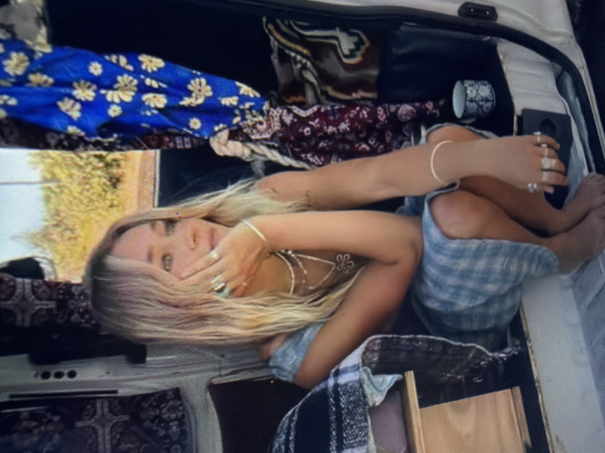 The image displays a young female, fair-skinned with long, wavy blonde hair, seated inside what appears to be a recreational vehicle or camper van. She is positioned in profile, facing and looking towards the right side of the frame, with her left hand raised and fingertips gently touching her chin. Her fingernails exhibit a light blue polish, and she wears a simple ring on her left pinky finger. Around her neck are multiple necklaces, including a layered white beaded strand, a multi-strand silver chain, and a silver necklace featuring a butterfly pendant. A small, dark tattoo is visible on her upper left arm. She wears a light blue and white checkered dress or top. Her right arm is visible resting, adorned with a thin white bracelet on her wrist and several silver rings on her thumb, index, middle, and ring fingers. Her bare feet rest on a light-colored surface at the bottom of the image.
The interior of the vehicle has light-colored walls. To the female's left, a dark, patterned blanket is draped over a wooden storage unit. Above her, a panel with three black circular controls is visible on the wall. To her right, a vibrant blue fabric with yellow floral patterns hangs, alongside a maroon fabric featuring lighter floral designs. A brown and gold patterned fabric is also visible on the right. A white ceramic mug with blue patterns rests on a dark surface to her lower right. The vehicle's white door frame is visible on the far right. Through an opening behind her, a blurred outdoor background shows green and yellow foliage, indicative of a natural or rural environment. There is no visual information within the image to identify the location as the city of Shumen, Bulgaria.