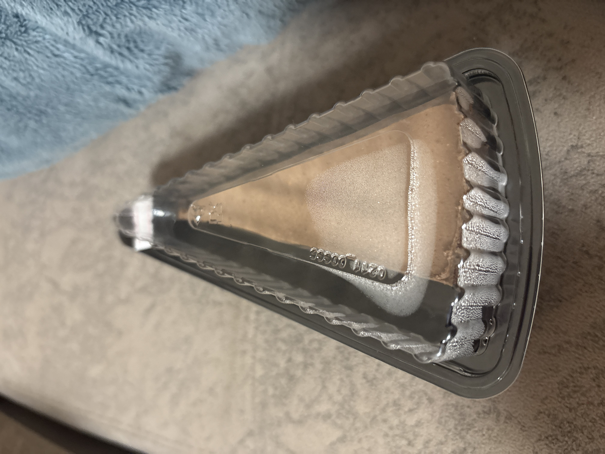 A triangular slice of cheesecake in a clear plastic container is placed on a beige, textured surface. The cheesecake appears to be light brown and has condensation on its surface and the inside of the container. A blue, fuzzy blanket is visible in the upper right corner of the image. The image does not provide any information about the location in the city of Kazan, Russia.