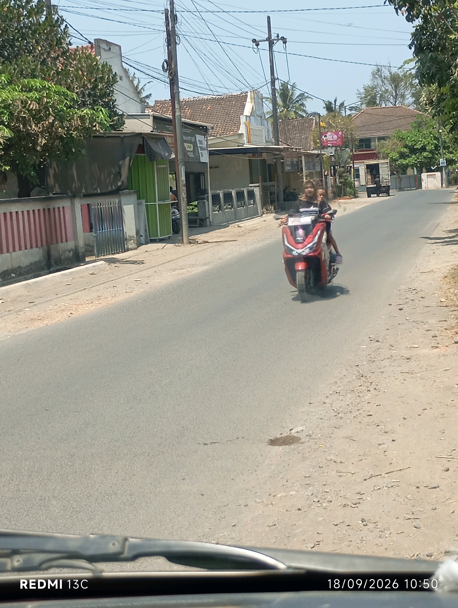 In this eye-level, medium shot, a red scooter with two occupants is traveling down a two-lane asphalt road in Sumberrejo, Indonesia. The rider, a female with long, dark hair, is wearing a dark top with white stripes on the sleeves. The passenger, who appears to be a child, is wearing a dark top and light-colored bottoms. They are traveling on the right side of the road, which is lined with buildings and vegetation. On the left side of the road, there is a pink and red striped fence, a building with a green facade, and other structures. On the right side of the road, there are more buildings, trees, and a utility pole. The sky is clear and blue, indicating a sunny day. The timestamp in the bottom right corner reads "18/09/2026 10:50".