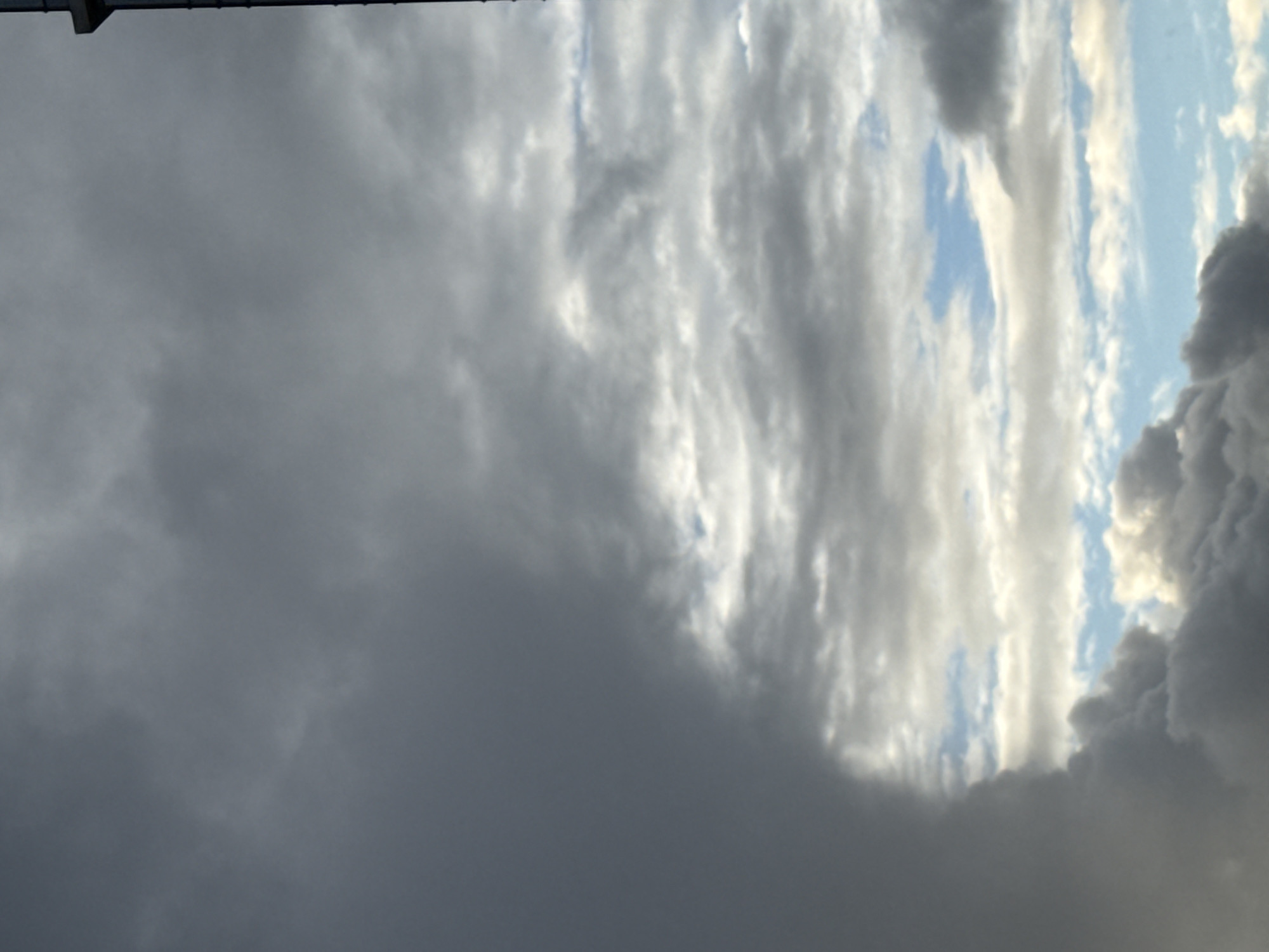 The image shows a sky filled with various types of clouds, likely taken in Khimki, Russia. Some clouds are dark and heavy, suggesting a possible storm, while others are lighter and more diffuse, allowing patches of blue sky to peek through. The overall scene conveys a sense of unsettled weather conditions. The image is rotated 90 degrees counterclockwise, so the top of the image is actually the left side of the frame in reality. In the upper left, there appears to be some dark metal object attached to the ceiling or roof.