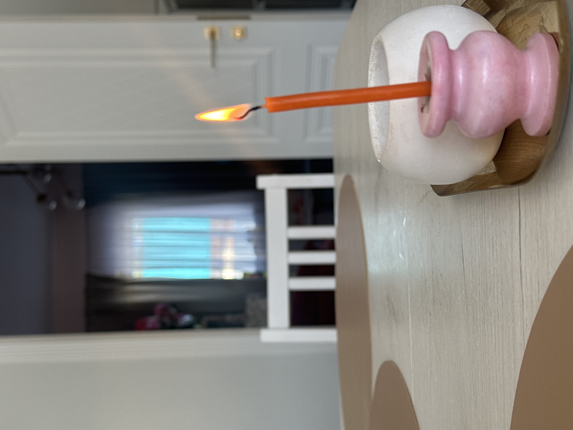 A lit orange candle is held in a white and pink decorative candle holder sitting on a wooden surface. A partially visible white door with a brass handle is in the background, with a darkened room behind it. A white chair back is also visible through the doorway. The light from a window is visible in the darkened room. The image is taken indoors. There is no indication of the location being Kharnavy, Russia.