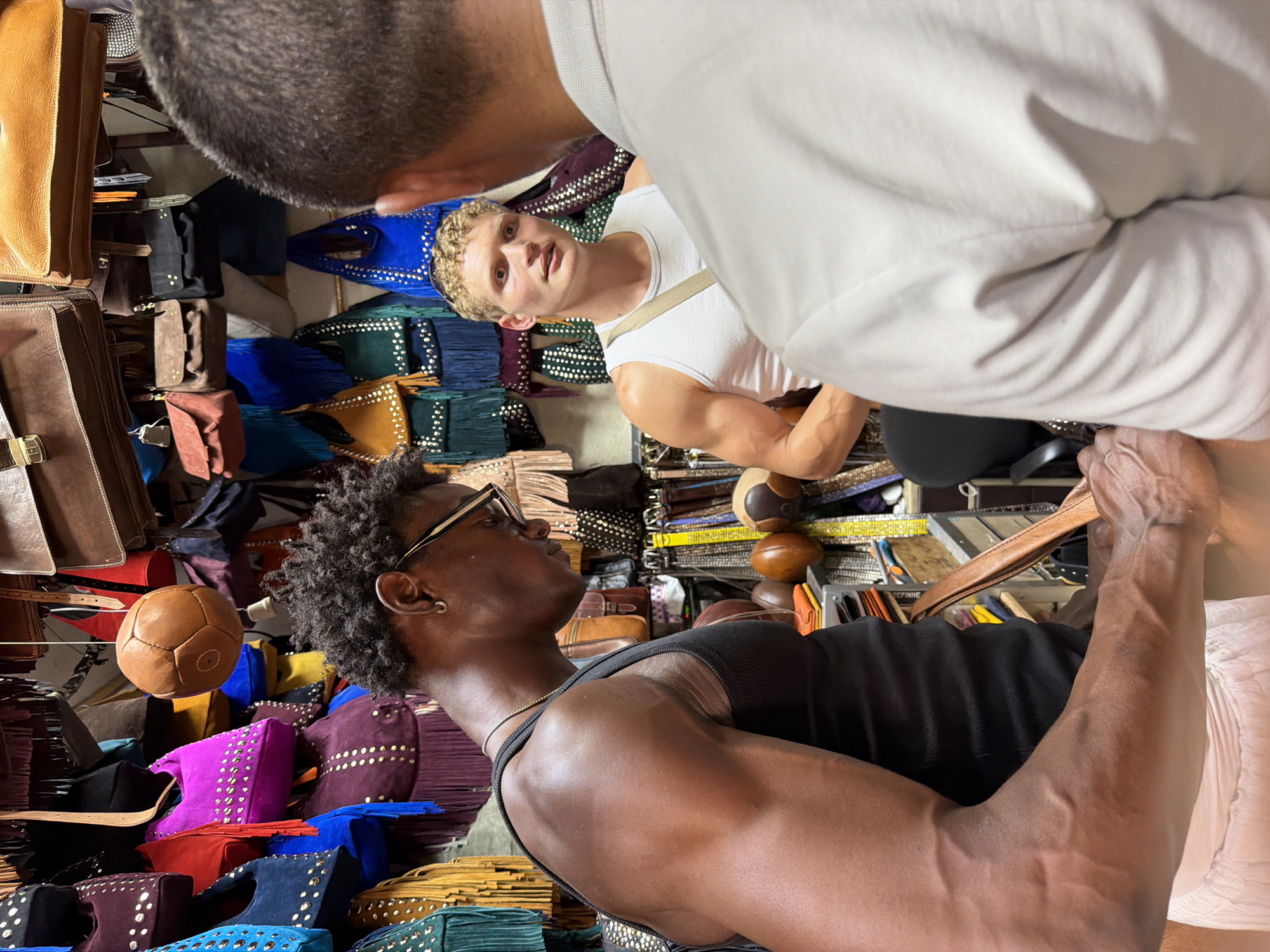 The image depicts an indoor market stall in Marrakesh, Morocco, likely within the Medina's souks, specializing in leather goods. Three individuals are present. In the foreground, a muscular Black man with dark, textured hair and glasses is seen from the chest up, wearing a black tank top and a light-colored skirt. His focus is directed downwards, as if examining an item he is holding, which appears to be a brown leather strap. To his right, a fair-skinned man with blond, curly hair and a white tank top is looking towards the Black man. He has a light-colored strap crossing his chest. Partially visible in the foreground, facing away from the camera, is a third individual wearing a light-colored polo shirt, obscuring their face. The background is filled with an array of colorful leather bags, predominantly in shades of blue, purple, red, yellow, and brown, many adorned with studs and fringes. A round, brown, stitched leather object, resembling a small ball, hangs from the ceiling. The overall scene suggests a commercial interaction, possibly a negotiation or product inquiry, within a vibrant artisan marketplace.