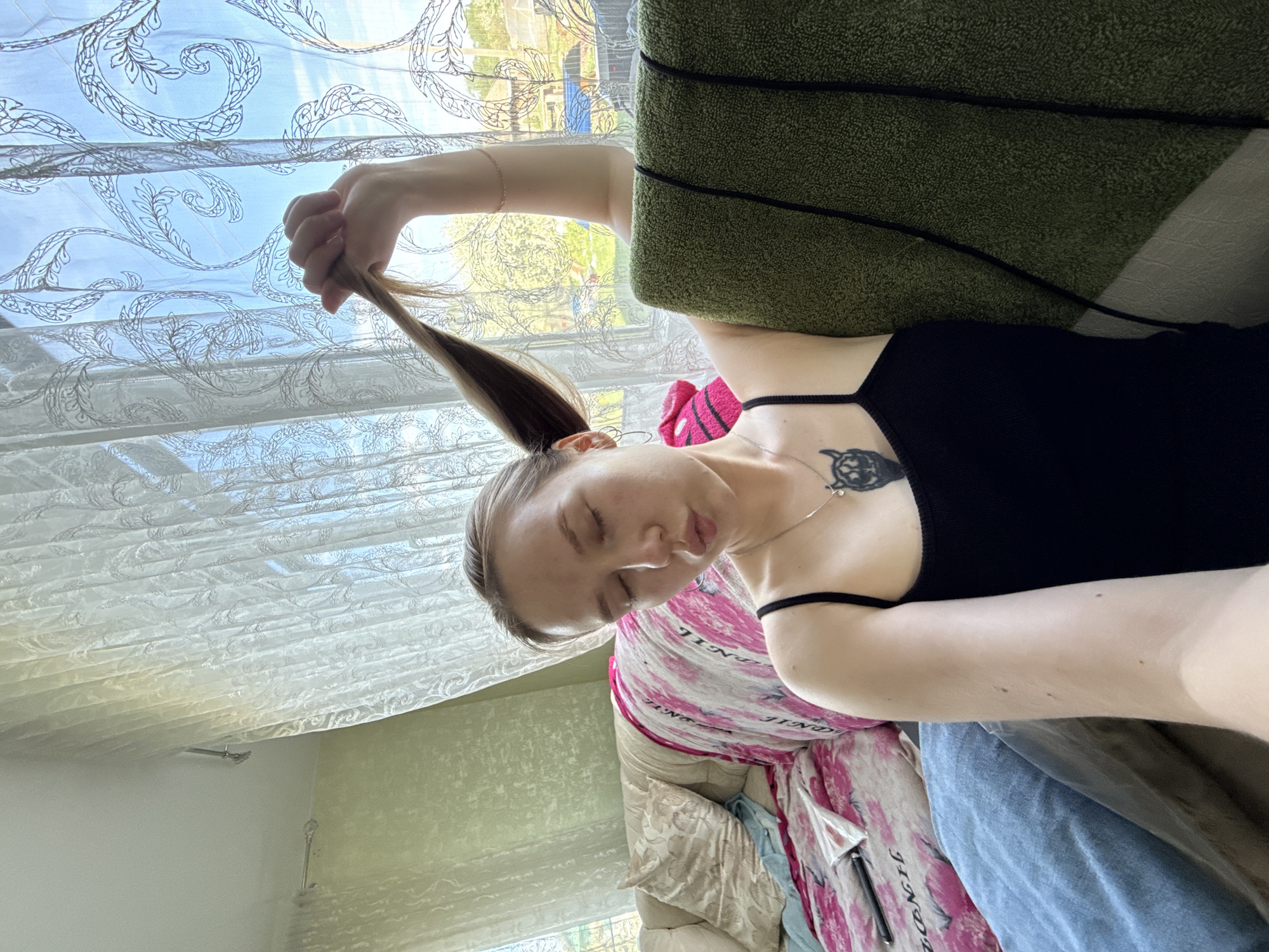 A woman is seated indoors in a room with a window, holding a section of her hair in her left hand. She is wearing a black tank top and a pink patterned shirt. A green object is positioned near her head. The room contains curtains, and a glimpse of the outside shows trees and buildings. The location is Unknown, Russia.