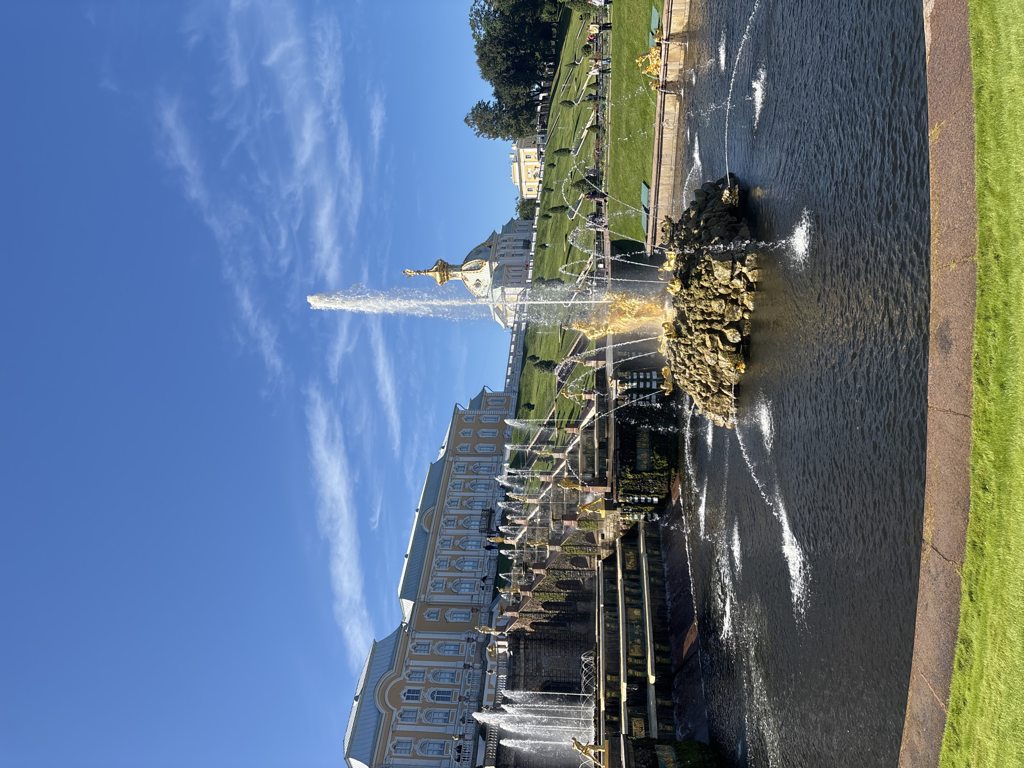 The image depicts the Grand Cascade at Peterhof Palace, a monumental fountain complex located in Peterhof, Russia, situated on the southern shore of the Gulf of Finland. In the foreground, a dark, reflective pool of water contains several spouting fountains. A large, central fountain rises from a rocky, gold-accented structure, with water dramatically spraying upwards and outwards. This central fountain is flanked by smaller fountains and decorative golden sculptures.
Behind the main fountain, a series of tiered terraces and staircases descend a grassy hill. Water flows down these terraces through numerous small waterfalls and cascades, feeding into the lower pool. Golden sculptures of mythological figures and sea creatures adorn the edges of the terraces and pools.
Further up the hill, the grand yellow palace of Peterhof Palace is visible, with its distinctive light yellow facade, white detailing, and numerous windows. To the right of the palace, a smaller, domed building with a golden cupola stands prominently on the elevated landscape. The entire area is meticulously landscaped with lush green lawns, neatly trimmed hedges, and scattered trees. The sky above is a vibrant, clear blue, with wisps of white clouds. Several people are scattered throughout the scene, walking on the paths and observing the fountains and palace. The lighting suggests a bright, sunny day.