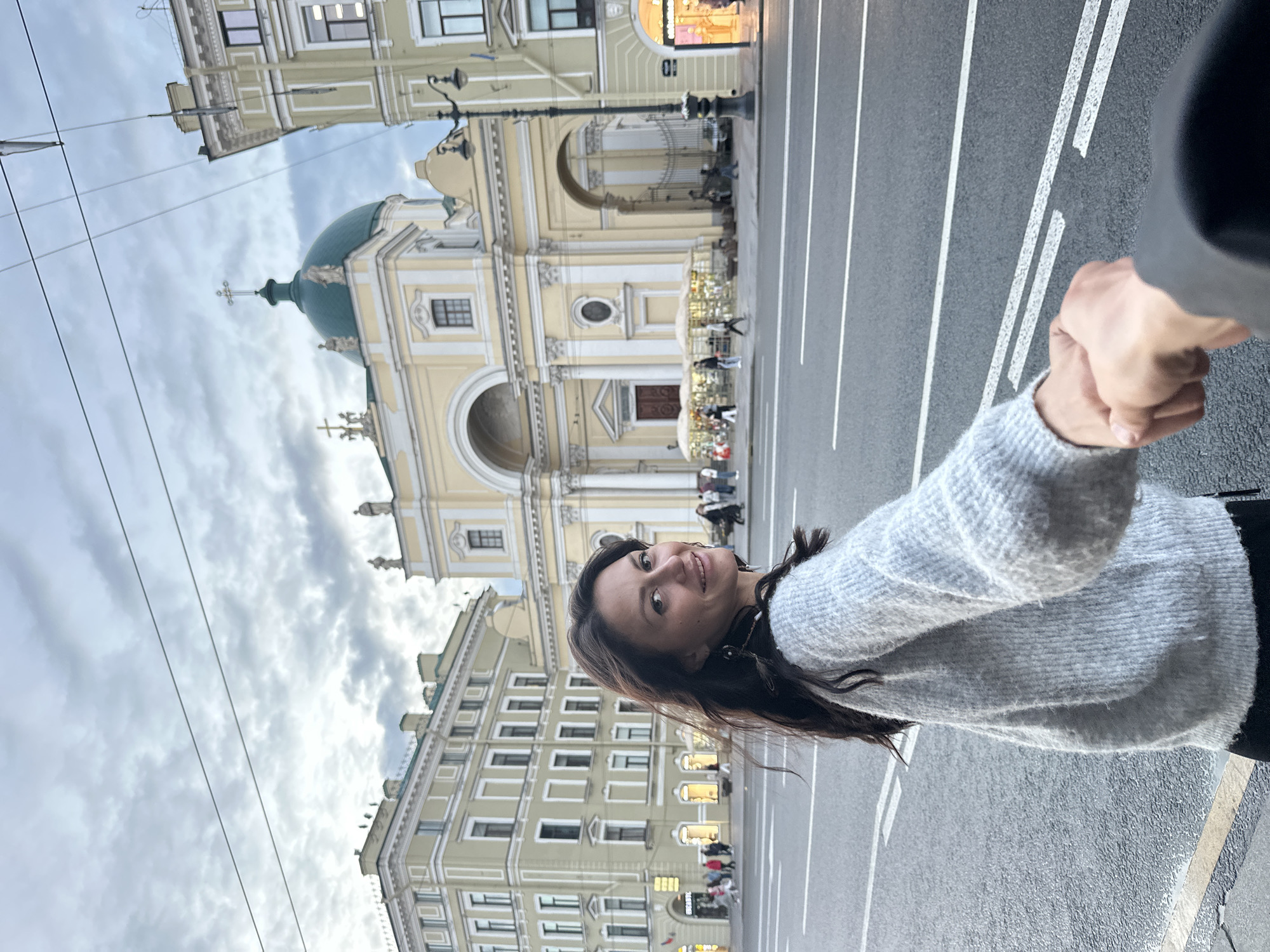 A woman, with long dark hair and a light gray sweater, smiles over her shoulder as she holds hands with an unseen person. She is standing on a city street in Saint Petersburg, Russia, with a large, pale yellow building with a green dome in the background. The building appears to be a church or a historically significant structure. The street in front of the building is wide with multiple lanes and white markings. The sky is overcast with clouds.