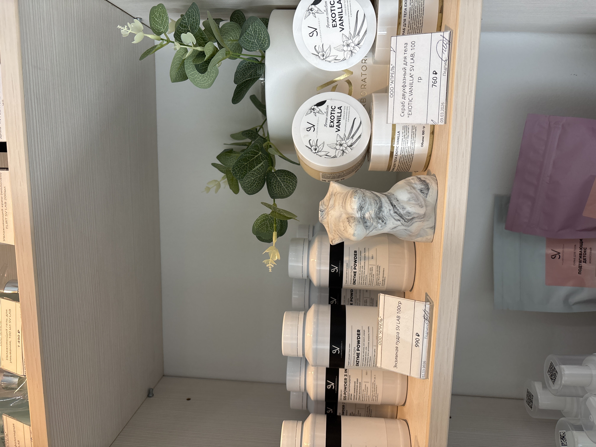 The image shows a shelf in a retail store, likely a beauty or cosmetics shop, featuring various skincare products. The shelf is made of light-colored wood and is filled with white and black bottles and jars. In the foreground, several white bottles with black labels are arranged, some of which appear to be enzyme powder. To the right, there are two round jars with "EXOTIC VANILLA" branding. A decorative bust of a woman's torso, made of a marbled material, is placed between the bottles and jars. In the background, a white pot with a green plant is visible. Various price tags and product information labels are attached to the items. The overall lighting suggests an indoor setting with artificial lighting. There are no people visible in the image. The environment appears clean and organized, typical of a retail display.