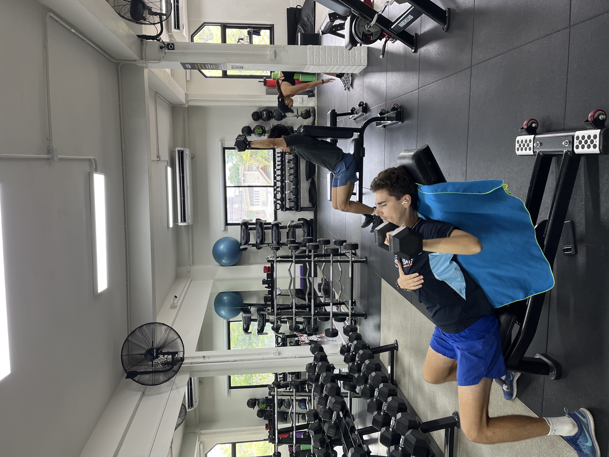 The image shows the interior of a gym with multiple people engaged in various exercises. In the foreground, a young man with short brown hair is performing a dumbbell exercise while lying on a weight bench, with a blue towel draped over the bench. He is wearing a dark t-shirt, blue shorts, white socks, and blue athletic shoes. Another young man in the background is also exercising with dumbbells, holding them with his arms extended. Further in the background, a woman is seen performing an exercise on a yoga mat. The gym appears well-equipped with various weights, dumbbells, barbells, and exercise machines. Large fans are mounted on the wall, and fluorescent lights illuminate the space. Windows with a view of trees and buildings are visible in the background, suggesting an urban setting. The overall mood seems to be one of focused exertion. The gym has a modern, clean aesthetic with grey flooring and white walls.