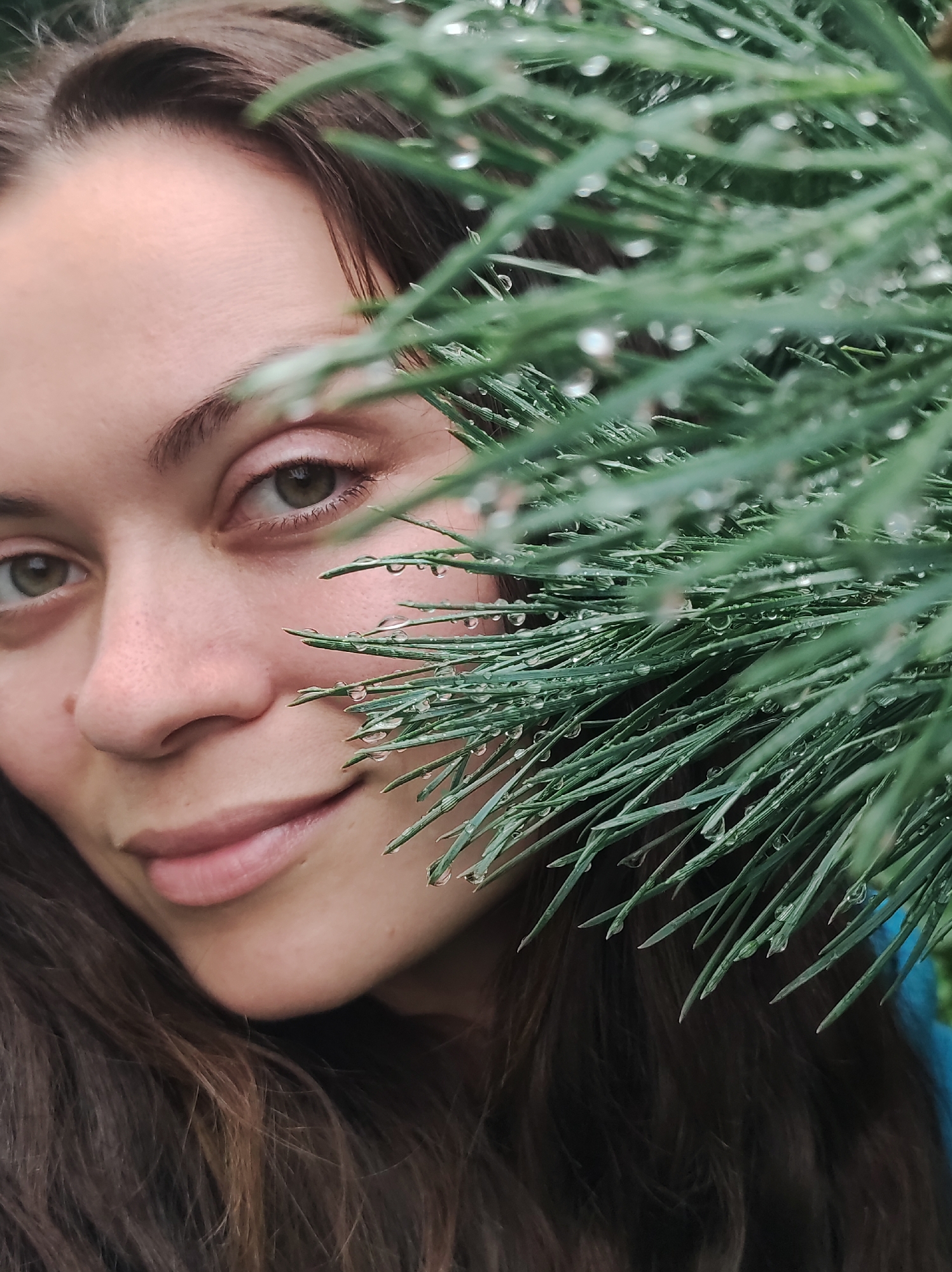A close-up, eye-level, outdoor shot features a woman's face partially obscured by a branch of evergreen foliage with water droplets. The woman's left eye, eyebrow, and part of her nose and mouth are visible. Her eyes are a light green or hazel color, and her skin is fair with some freckles. The foliage is green and appears to be pine needles or a similar conifer. The background is out of focus, but suggests an outdoor environment. The image is likely taken in the city of Fokino, Russia, due to the provided context. The woman is looking towards the viewer with a slight smile. The lighting is soft, suggesting overcast conditions or shade.