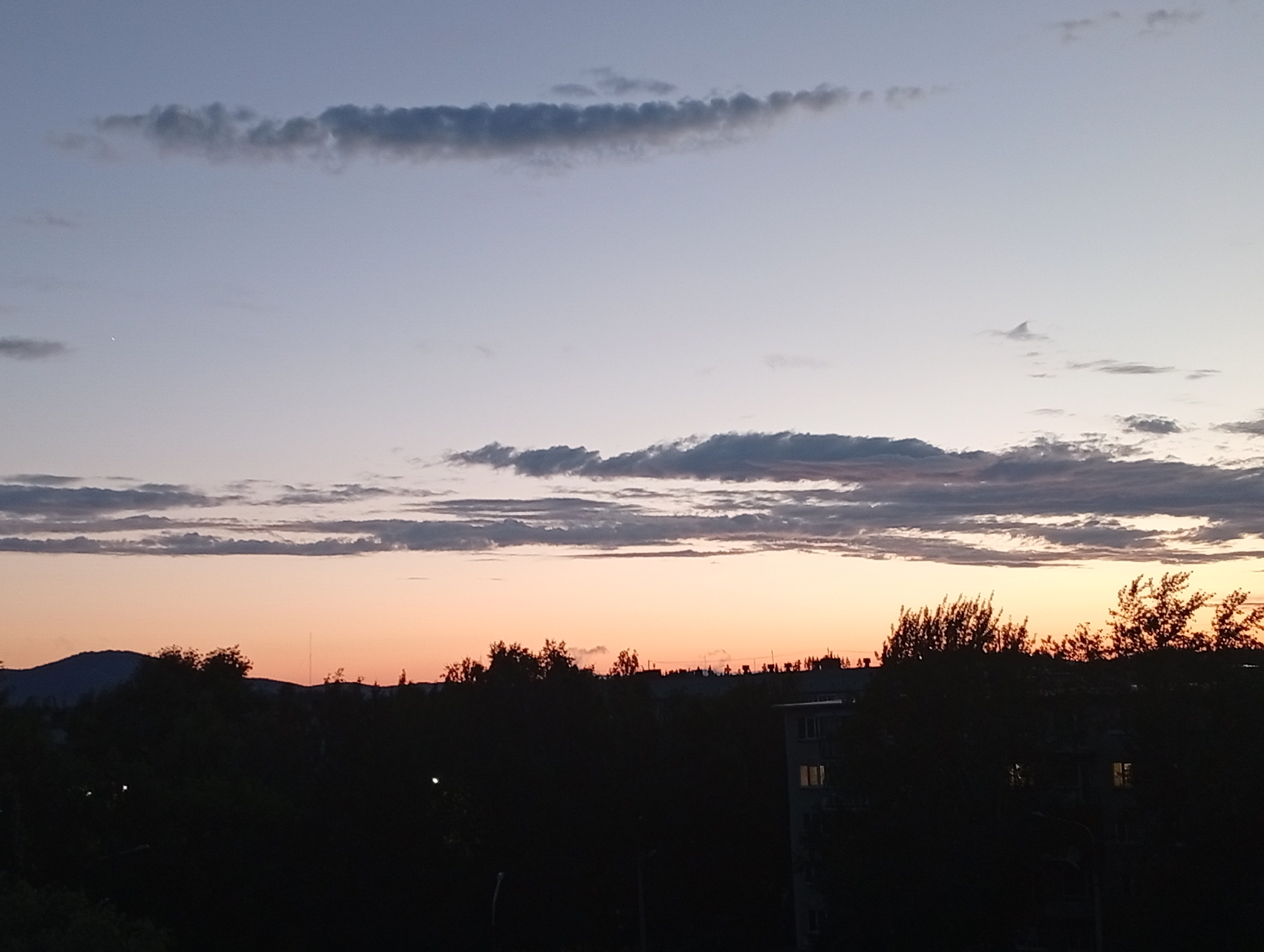 This media file depicts a twilight scene over Kyshtym, Russia. The sky transitions from a pale blue at the top to a warm peach and orange hue near the horizon, indicating either sunrise or sunset. A few wispy clouds are scattered across the sky, with some appearing as thin, elongated streaks and others as more textured formations.
Silhouetted against the colorful sky is a dark landscape featuring rolling hills or mountains in the distance to the left. In the foreground, the dense foliage of trees and the rooftops of buildings are visible as dark shapes. Several structures, likely apartment buildings, are discernible, some with illuminated windows suggesting interior lights are on. The overall impression is of a quiet evening or early morning in a town, with the natural beauty of the sky contrasting with the urban elements. There are no people or discernible objects in the foreground, and no visible text. The atmosphere conveyed is serene and peaceful.