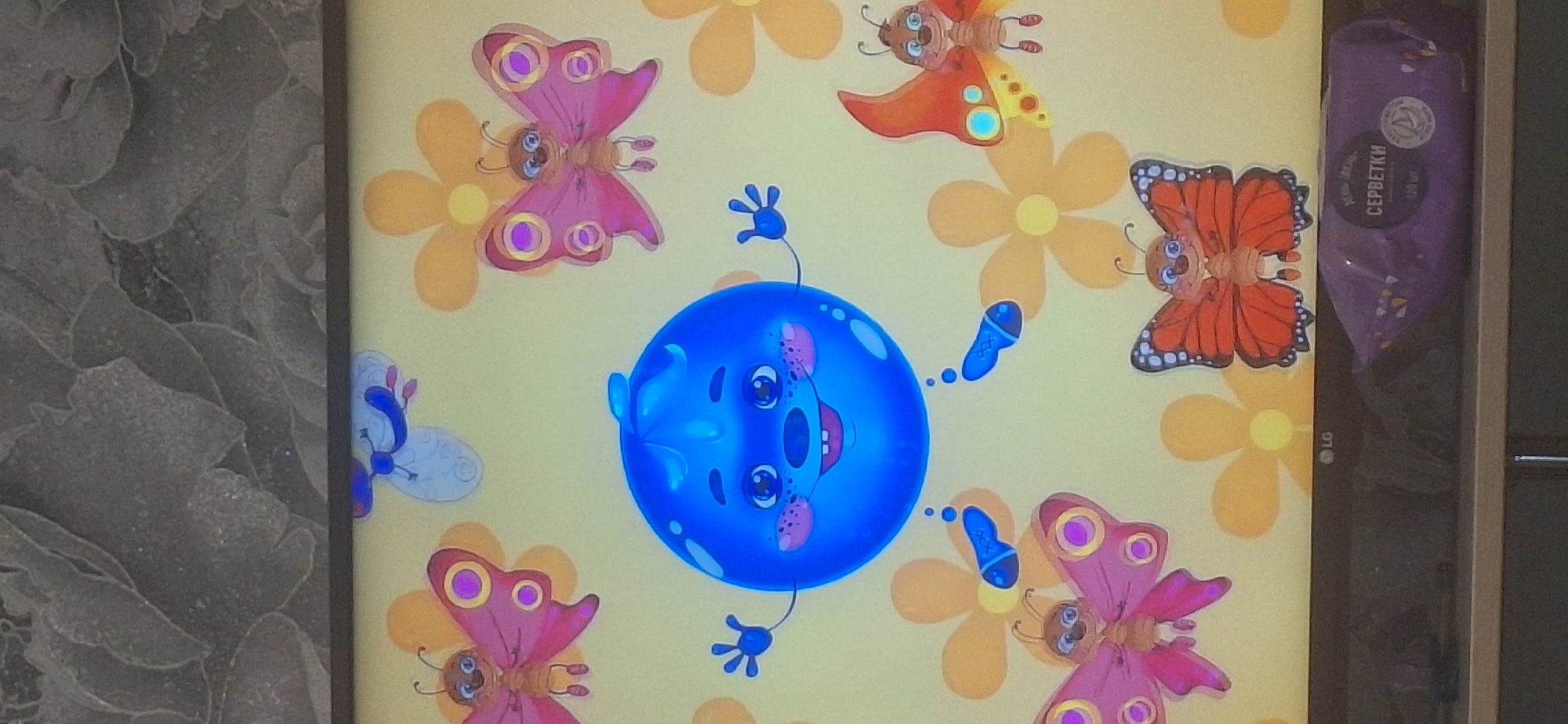 The image shows a television screen displaying a cartoon of a smiling blue blueberry character with arms and legs, surrounded by butterflies and flowers on a yellow background. The television is placed on a shelf, below which is a package of wet wipes. The LG logo is visible on the bottom of the television screen. The background of the image above the screen appears to be a wall with a textured wallpaper. There is no indication of the specific location within Bila Tserkva, Ukraine.