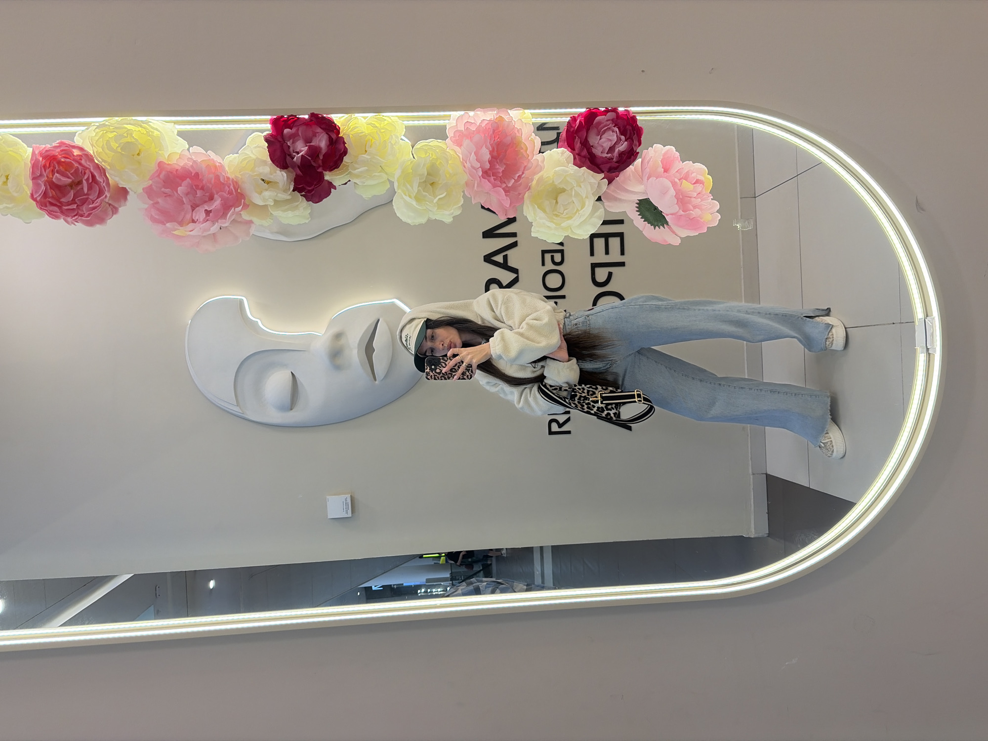 A young woman is taking a mirror selfie in what appears to be a retail store. She is wearing a cream-colored hooded sweatshirt, light blue wide-leg jeans, a baseball cap, and white sneakers. She is holding a phone with a leopard-print case and has a leopard-print crossbody bag. Behind her, a large mirror reflects the store's interior, which includes a white sculpture of a face and a string of artificial flowers in shades of pink and yellow. Text, possibly a store name, is visible on the wall in the reflection. The mirror has a bright LED light strip around its edge. The floor is tiled with black and white squares. The woman's reflection shows her standing on the tiled floor. The scene suggests a trendy shopping environment. There is no identifiable information to place this scene in Saint Petersburg, Russia.
