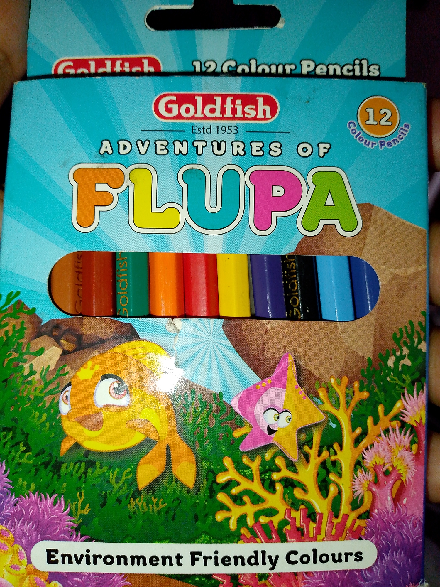 A product box for "Goldfish Adventures of Flupa" colored pencils is depicted, featuring a cartoon underwater scene. The box is primarily blue with stylized rays emanating from the top. The brand name "Goldfish" is prominent in red and white. Below this, "Adventures of Flupa" is written in large, colorful block letters. The phrase "12 Colour Pencils" is also visible. A circular emblem with the number "12" and "Colour Pencils" in a handwritten font is on the upper right. A cutout in the box reveals a set of twelve colored pencils, some with "Goldfish" printed on them. The lower portion of the box displays cartoon characters: a smiling orange fish on the left and a pink and yellow starfish on the right, set against a background of colorful coral and seaweed. The text "Environment Friendly Colours" is printed on a white label at the bottom of the box. The image appears to be a photograph taken in an indoor setting, likely a retail environment or a home. There are no identifiable landmarks or contextual clues to determine a specific city or location.