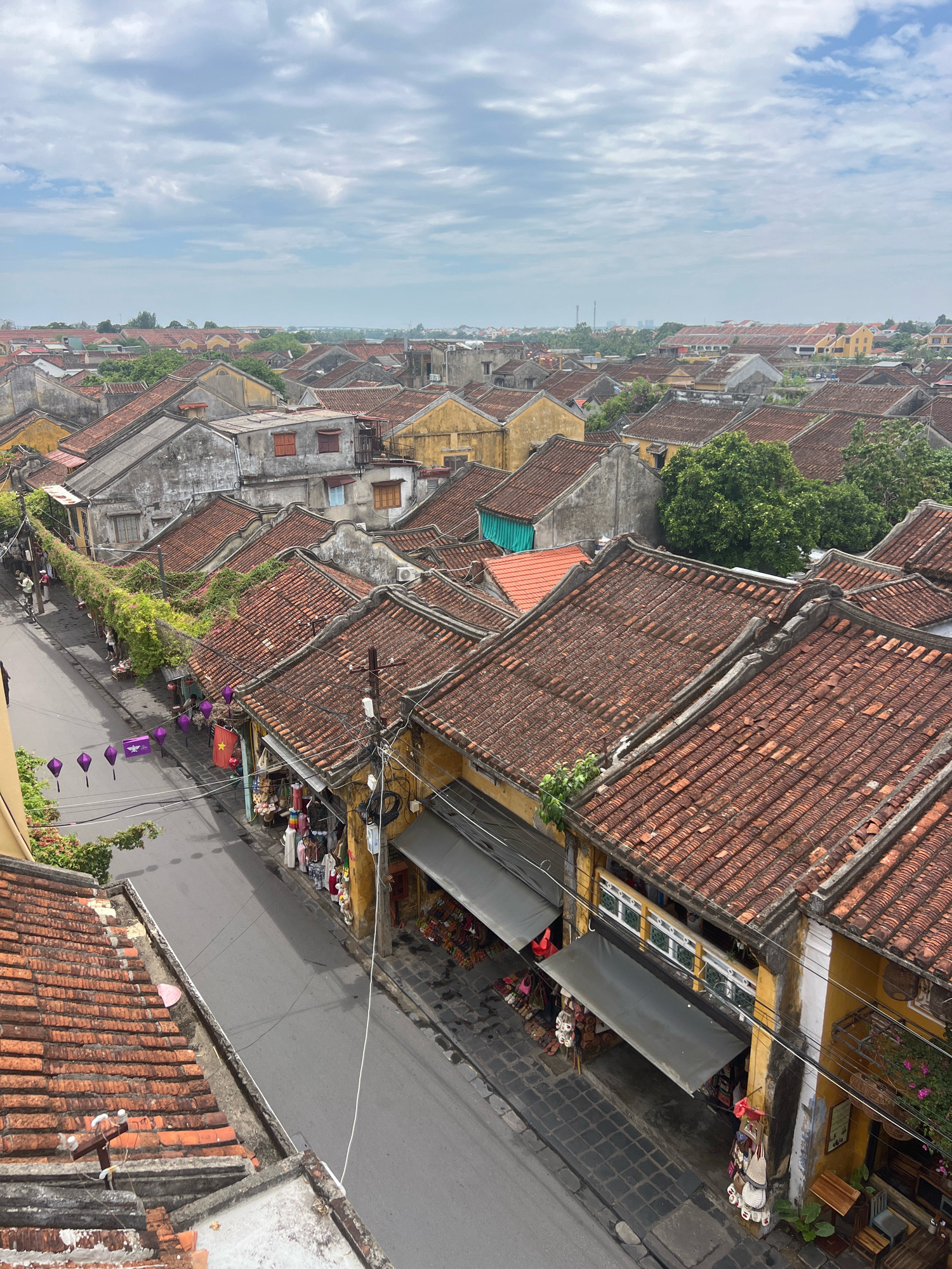 The image is an aerial view of the ancient town of Hoi An, Vietnam, specifically a street lined with traditional yellow buildings featuring terracotta tiled roofs. The street is paved with grey asphalt and bordered by stone sidewalks. On the left side of the street, a row of buildings with red tiled roofs and yellow walls are visible, some with greenery growing over them. Purple lanterns are strung across the street in several places. Several shops with awnings are set up along the right side of the street, displaying various goods such as bags, clothing, and footwear. A few people are walking on the sidewalks and a small Vietnamese flag is hanging from one of the buildings. The sky is cloudy, with patches of blue visible. The architecture is typical of Hoi An's historic district, characterized by its well-preserved wooden buildings and tiled roofs.