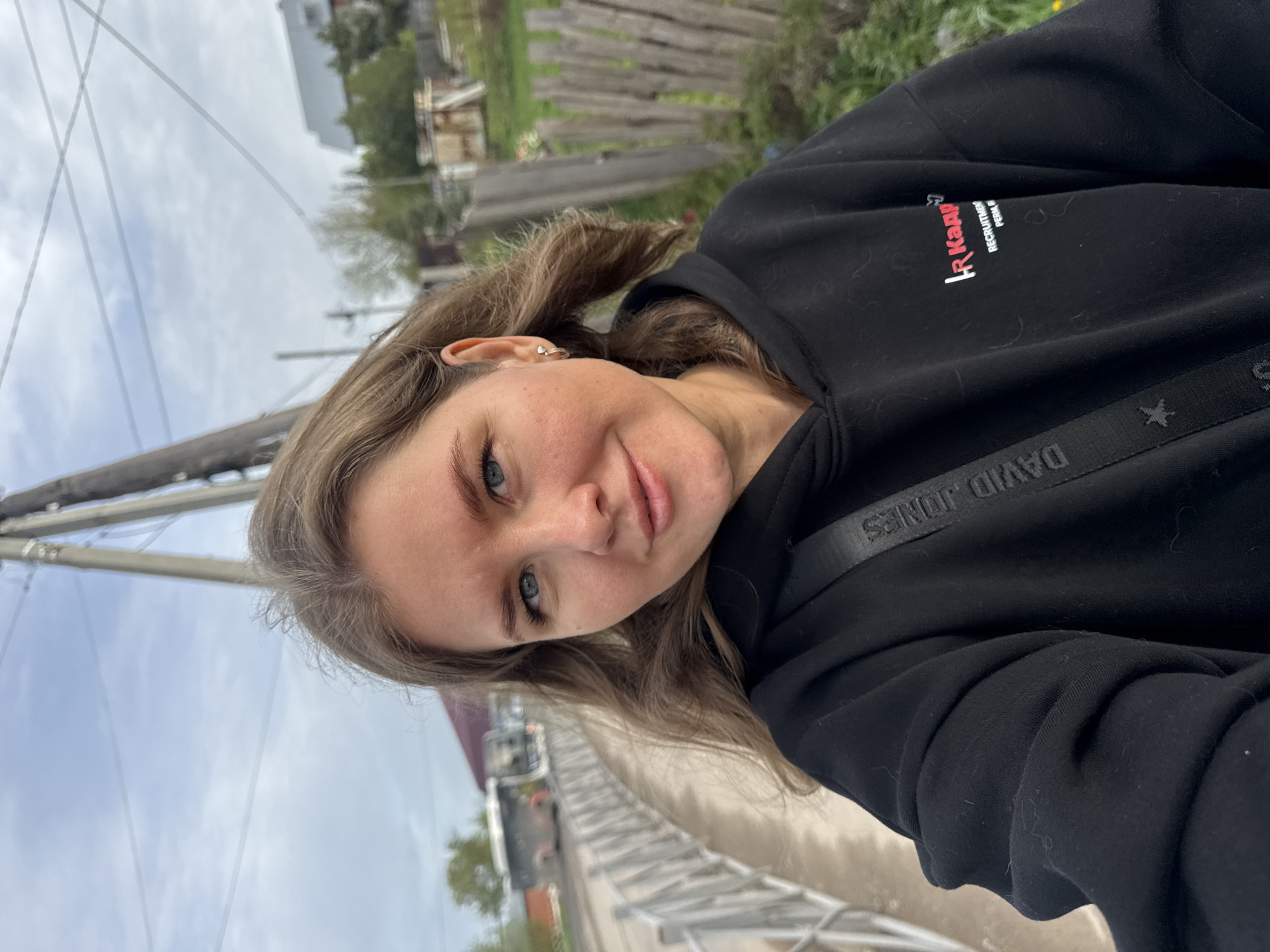 A woman with fair skin, light brown hair, and blue eyes is centered in a medium, angled shot. She is wearing a black hooded sweatshirt. The name "David Jones" is visible on a strap crossing her torso. Part of a building is visible in the background, along with a fence and green vegetation. The sky is overcast. Utility poles and wires are also visible. Based on the location in the image, the location is in Ферма, Russia.