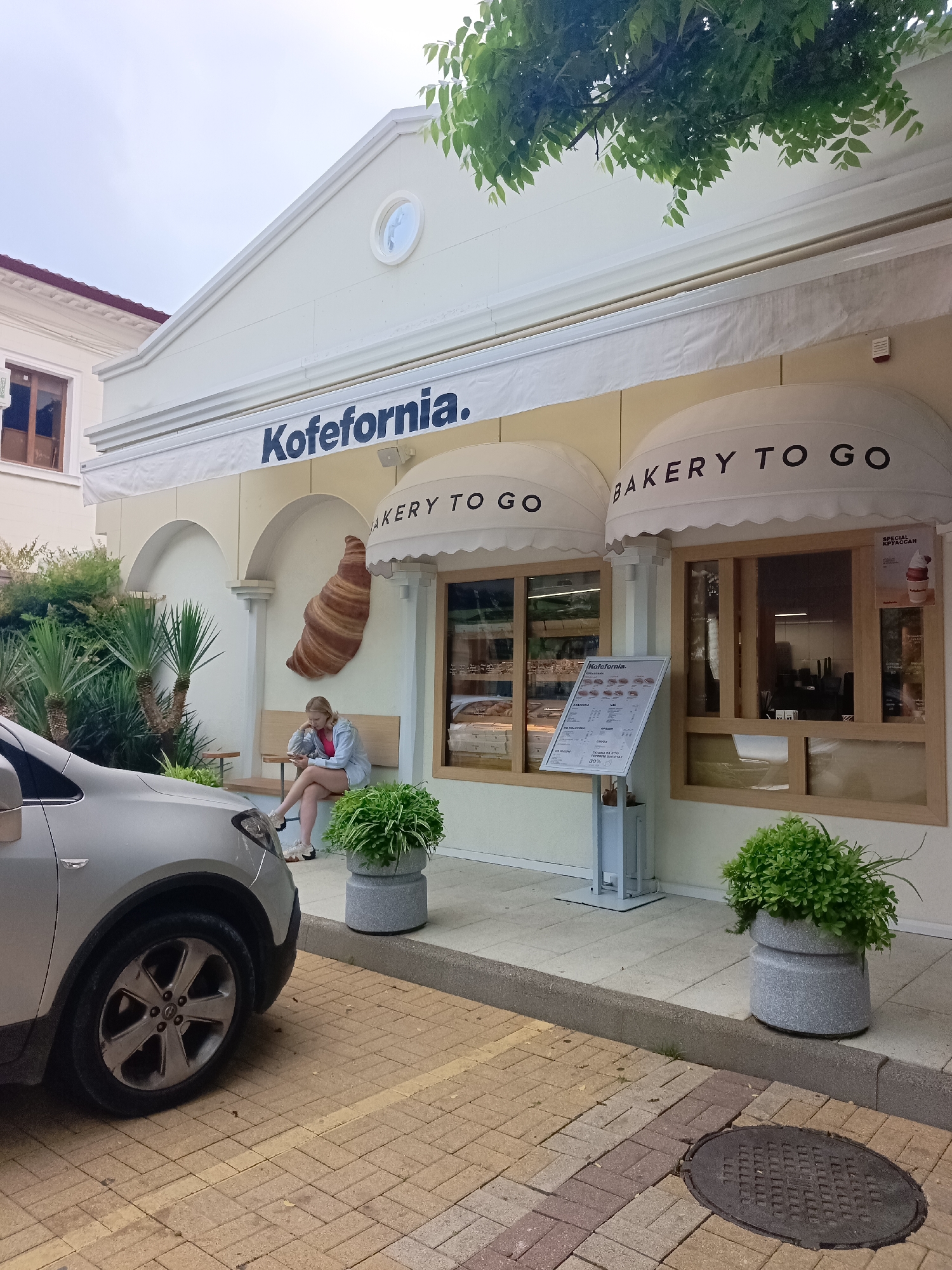 An outdoor shot shows a street-level view of a bakery storefront named "Kofefornia." The building has a white facade with light yellow accents around arched windows. Two white awnings extend over the windows, each with text reading "BAKERY TO GO." A large, brown, wall-mounted croissant decoration is visible on the left side of the building. In front of the bakery, a young blonde woman with light skin and a pink top is seated on a wooden bench, looking at her phone. She is wearing denim shorts and a light blue jacket. To the left of the woman and bench, there are some green palm-like plants. In the foreground, the front quarter of a silver car is visible, parked on a paver stone driveway. To the right of the car, two large, round, grey planters filled with lush green plants are positioned on the sidewalk. A metal manhole cover is located on the paver stones in the lower right corner. The sky is overcast. The style of architecture and the presence of palms suggest a warm climate, consistent with Sochi, Russia.