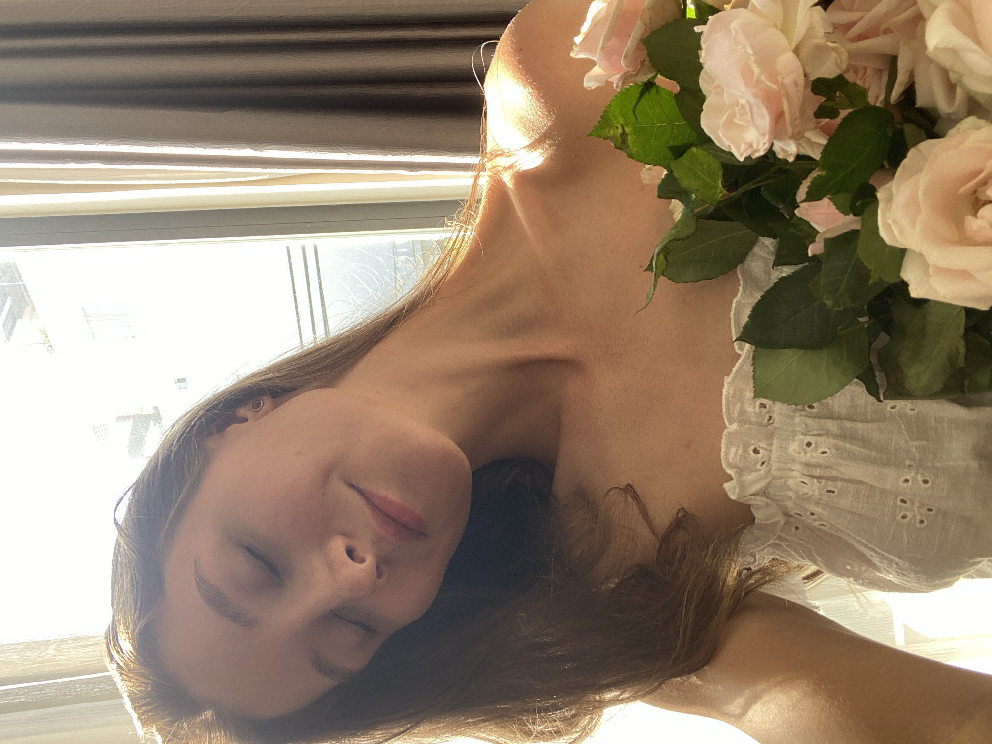 A young woman with her eyes closed and head tilted back, bathed in sunlight, holds a bouquet of pink roses. She is wearing a white, off-the-shoulder eyelet top. The background features a window with sheer curtains and drapes, with light streaming in. The scene suggests a peaceful, indoor setting, possibly a bedroom or living area, located in Bac Nha Trang Ward, Vietnam.