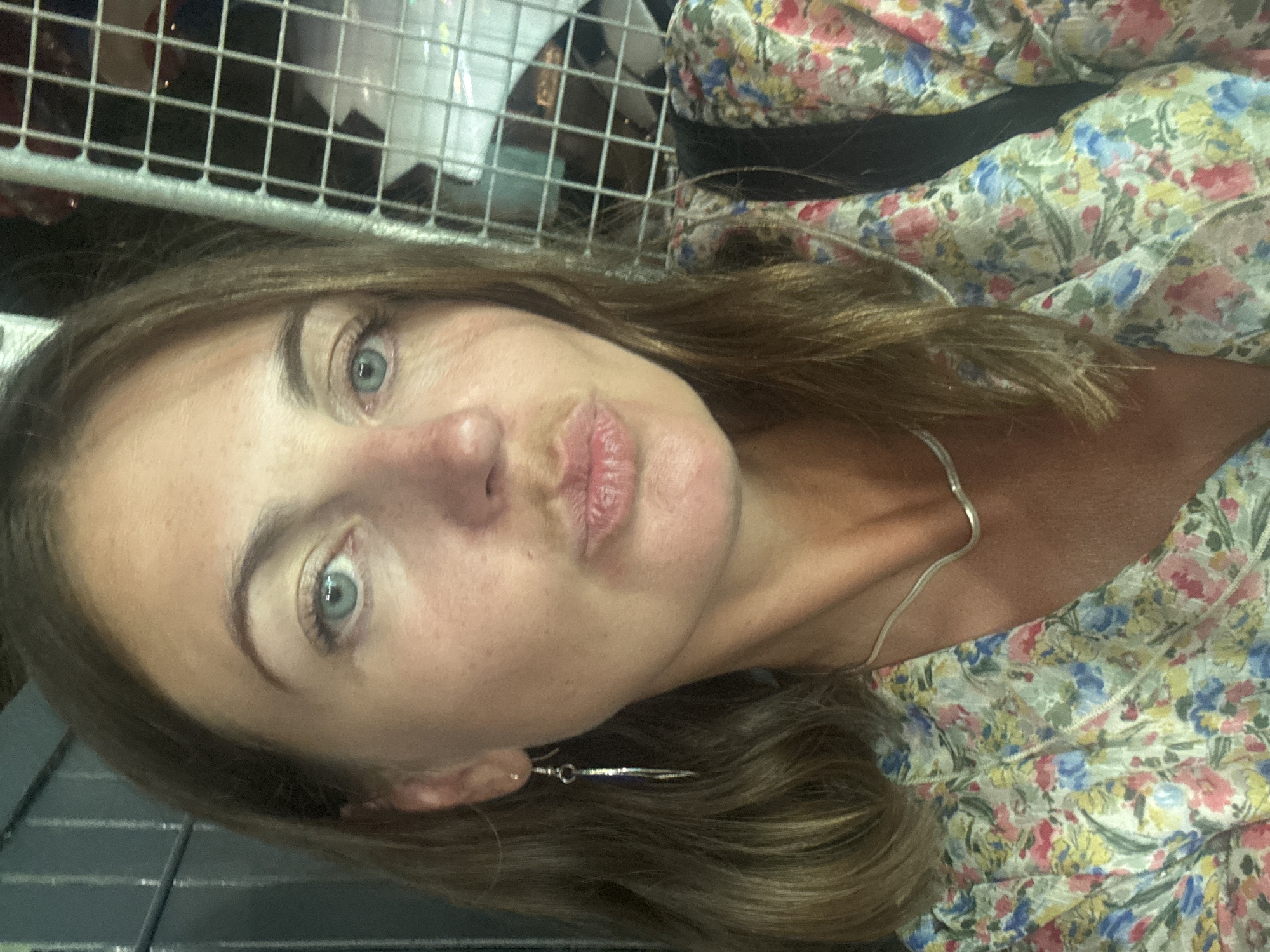 A woman with shoulder-length brown hair and blue eyes is posing for a selfie. She is wearing a floral print top and a silver necklace. She has her lips pursed in a kiss pose. The background appears to be a metal grate and possibly an indoor setting, with some blurred figures in the distance. The image was taken in Argelès-sur-Mer, France.
