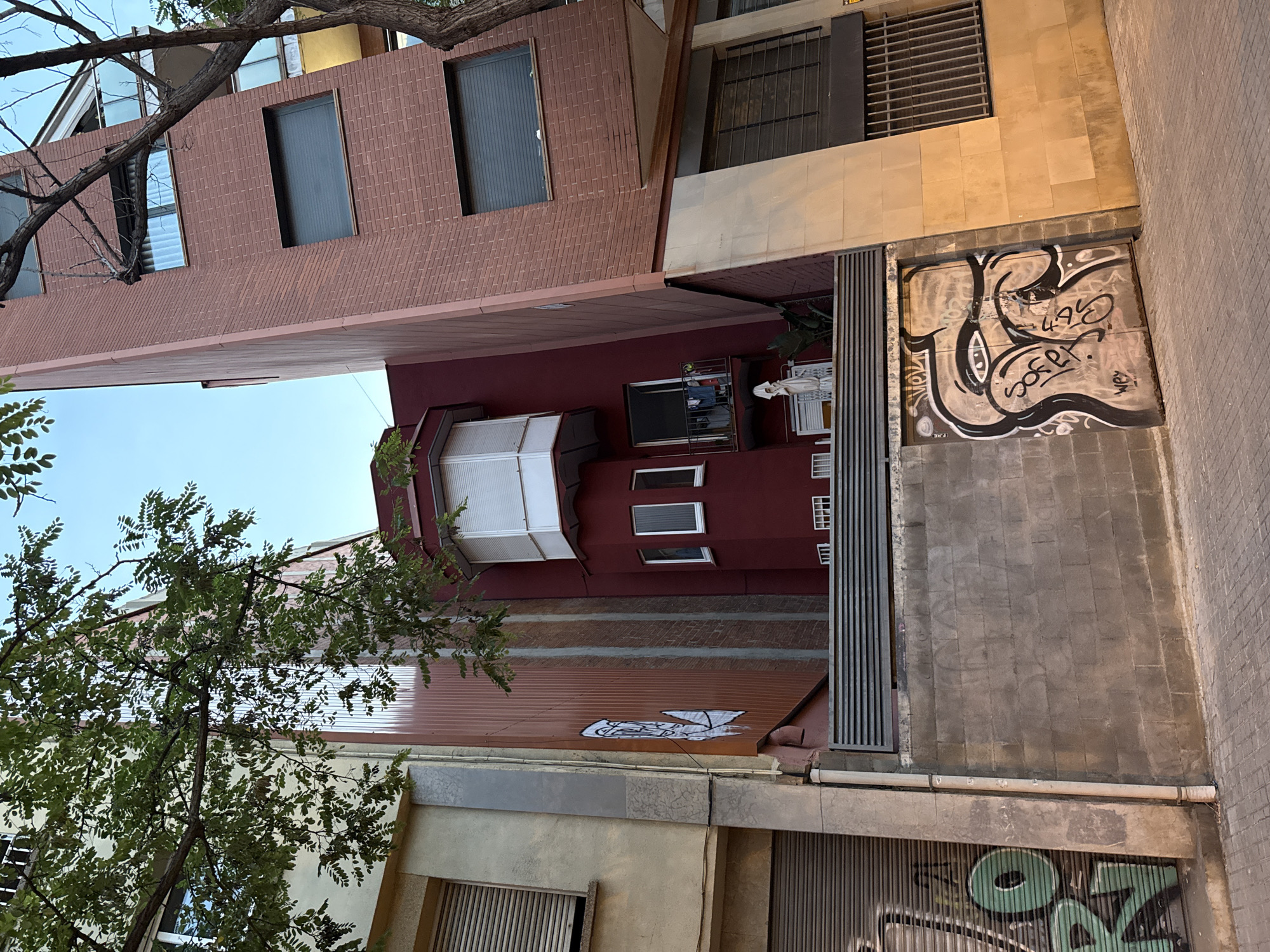 The image shows a narrow street in Barcelona, Spain, lined with buildings of varying architectural styles and colors. The buildings are densely packed, with some leaning towards each other, creating a sense of verticality and enclosure. Graffiti is visible on some of the lower walls and a metal gate. A tree with green leaves partially obscures the view of the buildings from the foreground. The sky is clear and blue, suggesting it is daytime. The overall impression is a vibrant urban environment with a mix of old and new architecture and elements of street art.