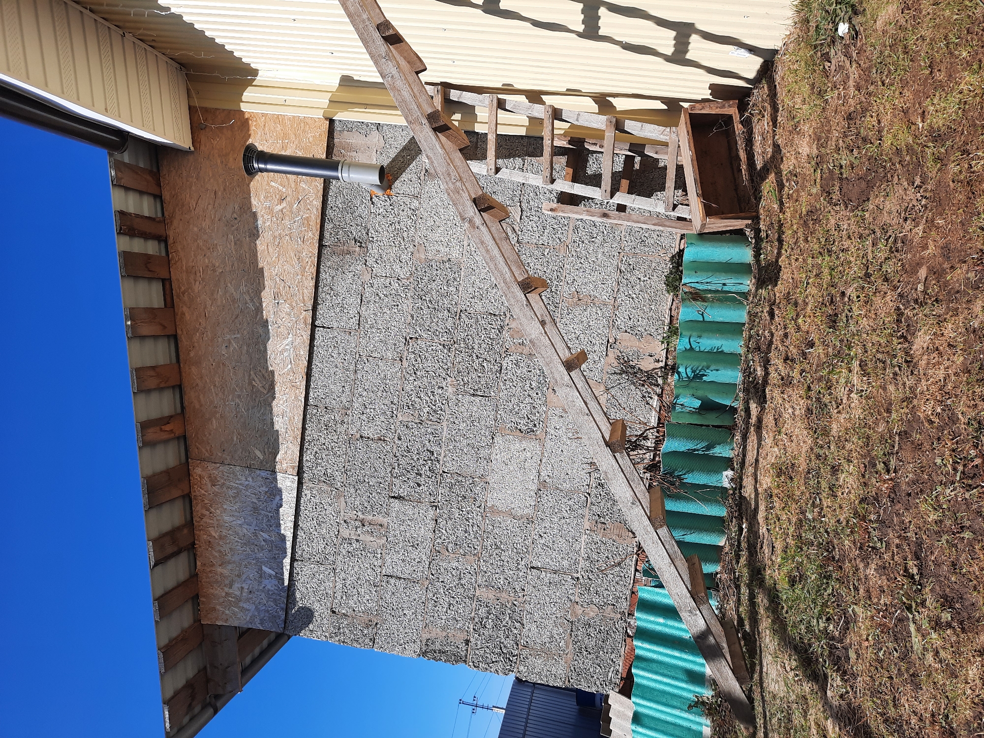 The image shows the exterior of a building in Krasnogorskoye, Russia. The building's siding consists of various materials including wood paneling and textured gray blocks. A section of the wall has a black pipe extending from it. An old wooden ladder is leaning against the wall. Adjacent to the building is a patch of dry grass and a stack of green corrugated sheets.