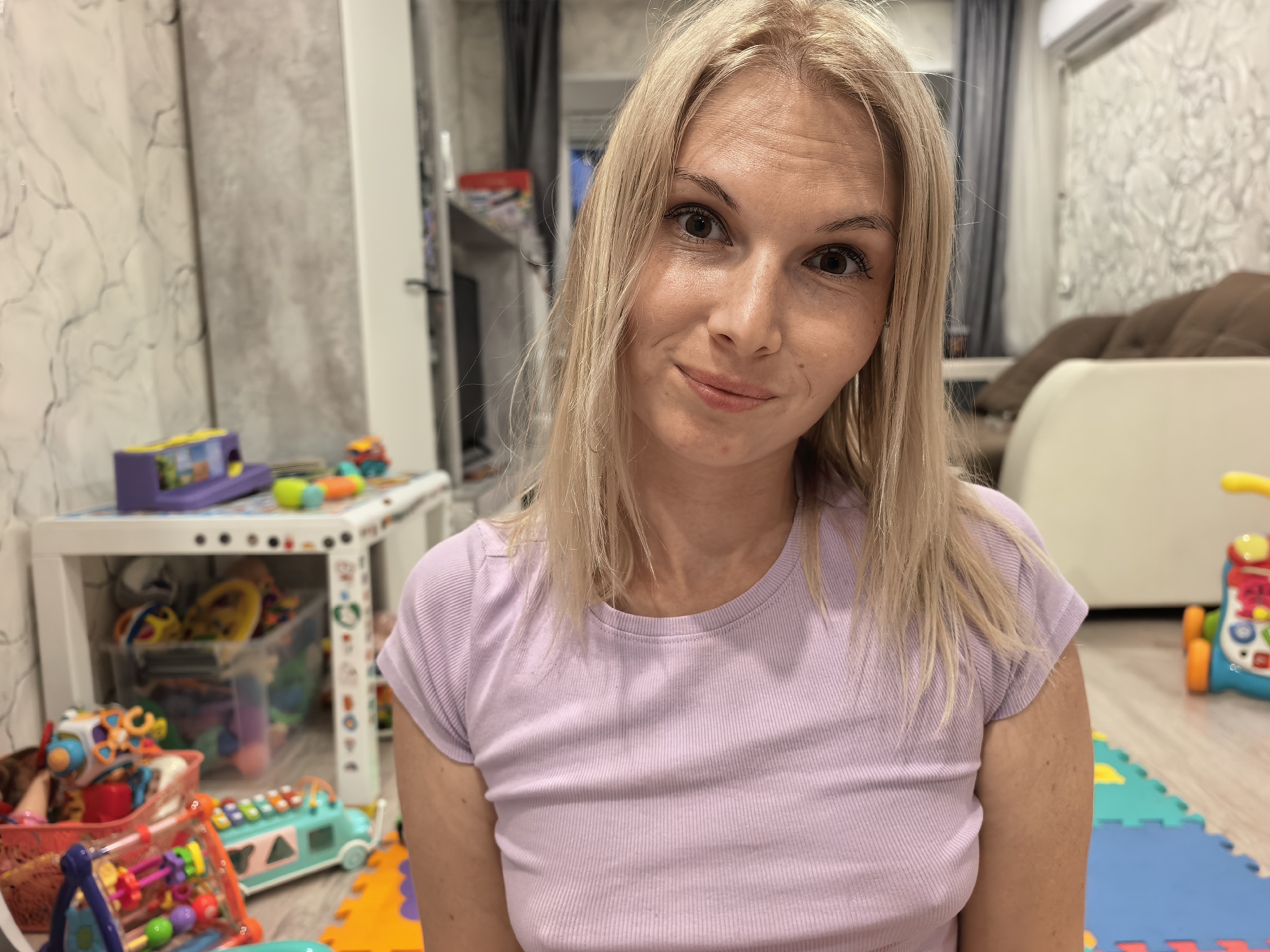 A woman with blonde hair is seated in a room filled with children's toys. She is wearing a light purple ribbed t-shirt and looking directly at the camera with a slight smile. The background shows a white shelf unit stocked with more toys, a wall with textured wallpaper, and a glimpse of a couch and air conditioning unit. The floor is covered with colorful interlocking foam mats. The scene appears to be a child's playroom or a living area adapted for children, likely in a residential setting.