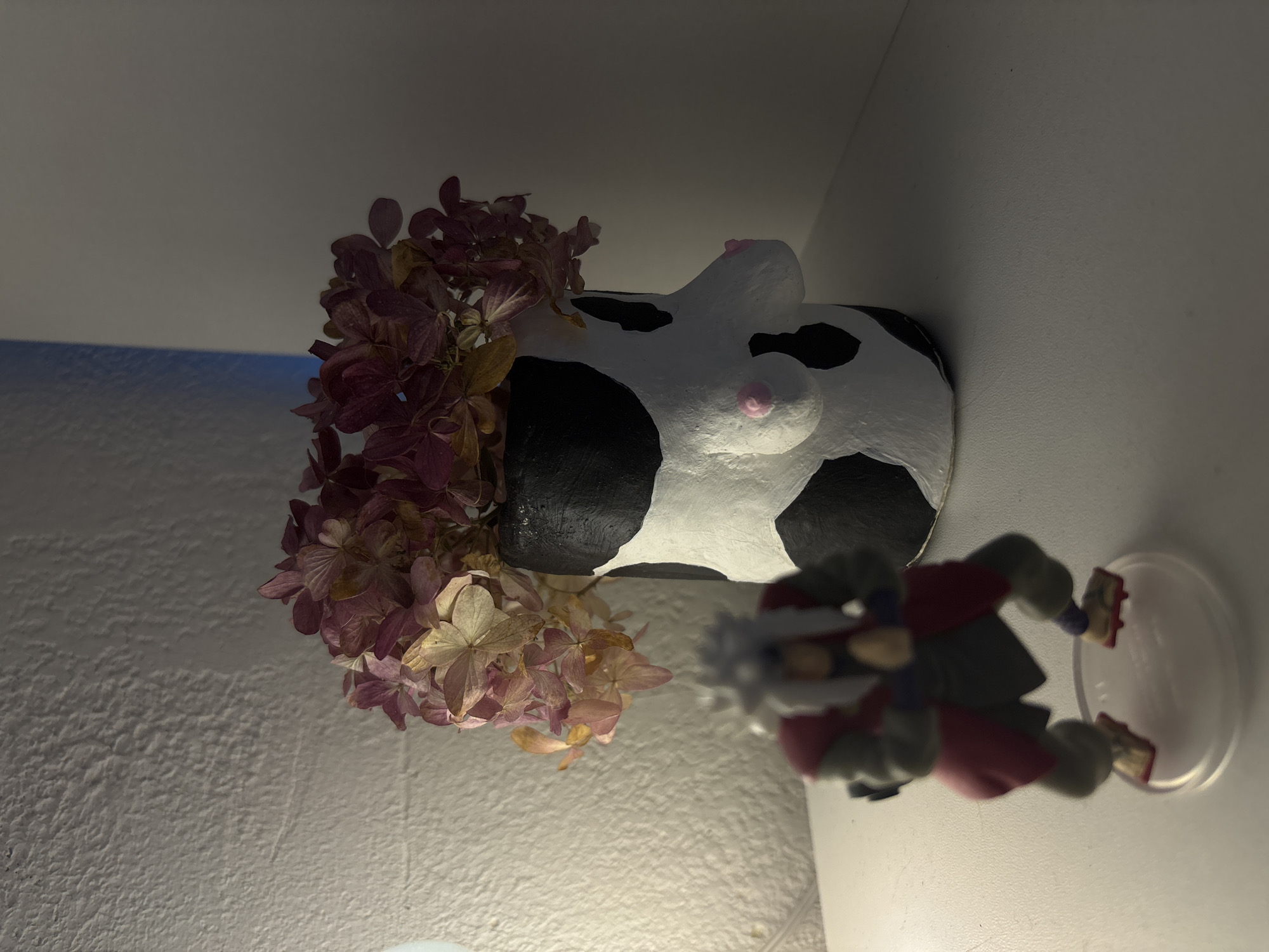 The image shows a small, white cow-shaped vase with black spots and pink protrusions, filled with dried pink and beige hydrangea flowers. Beside the vase is a small, blurred figurine of a creature in grey, red, and white colors, standing on a clear plastic base. The vase and figurine are positioned in the corner of a room with white walls and are illuminated by a warm, dim light source. The location is not discernible from the image.