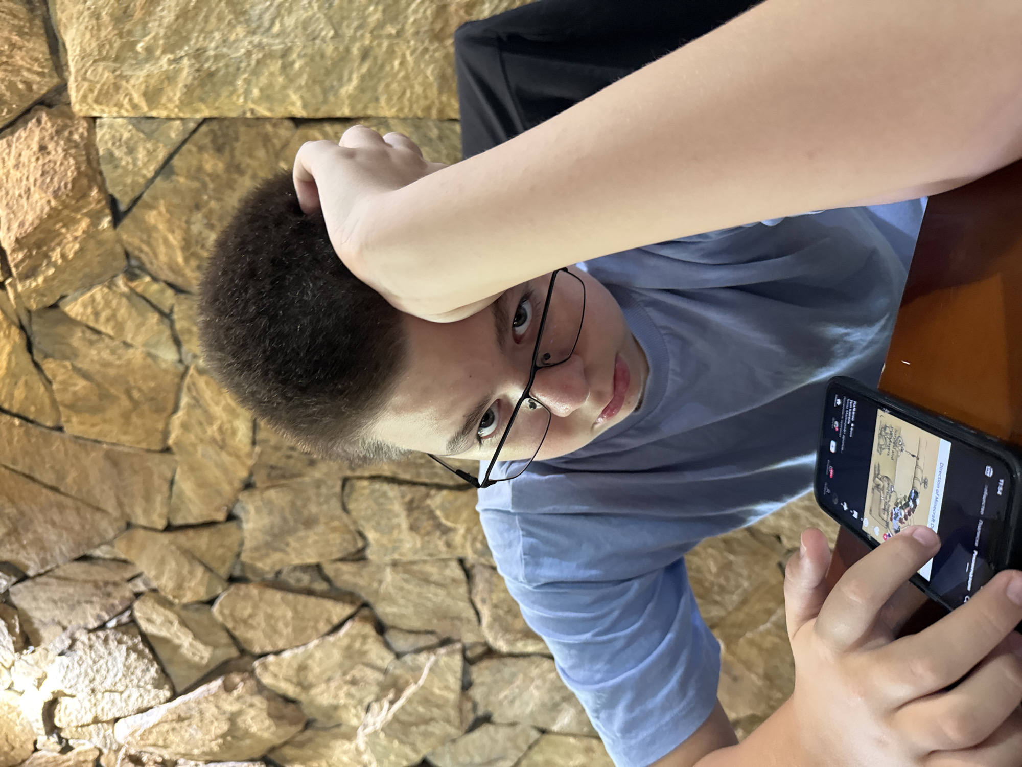 A boy wearing a light blue t-shirt and glasses is sitting at a table, looking at a smartphone. His left arm is resting on his head, and his right hand is holding the phone. The background features a stone wall. The scene is indoors.