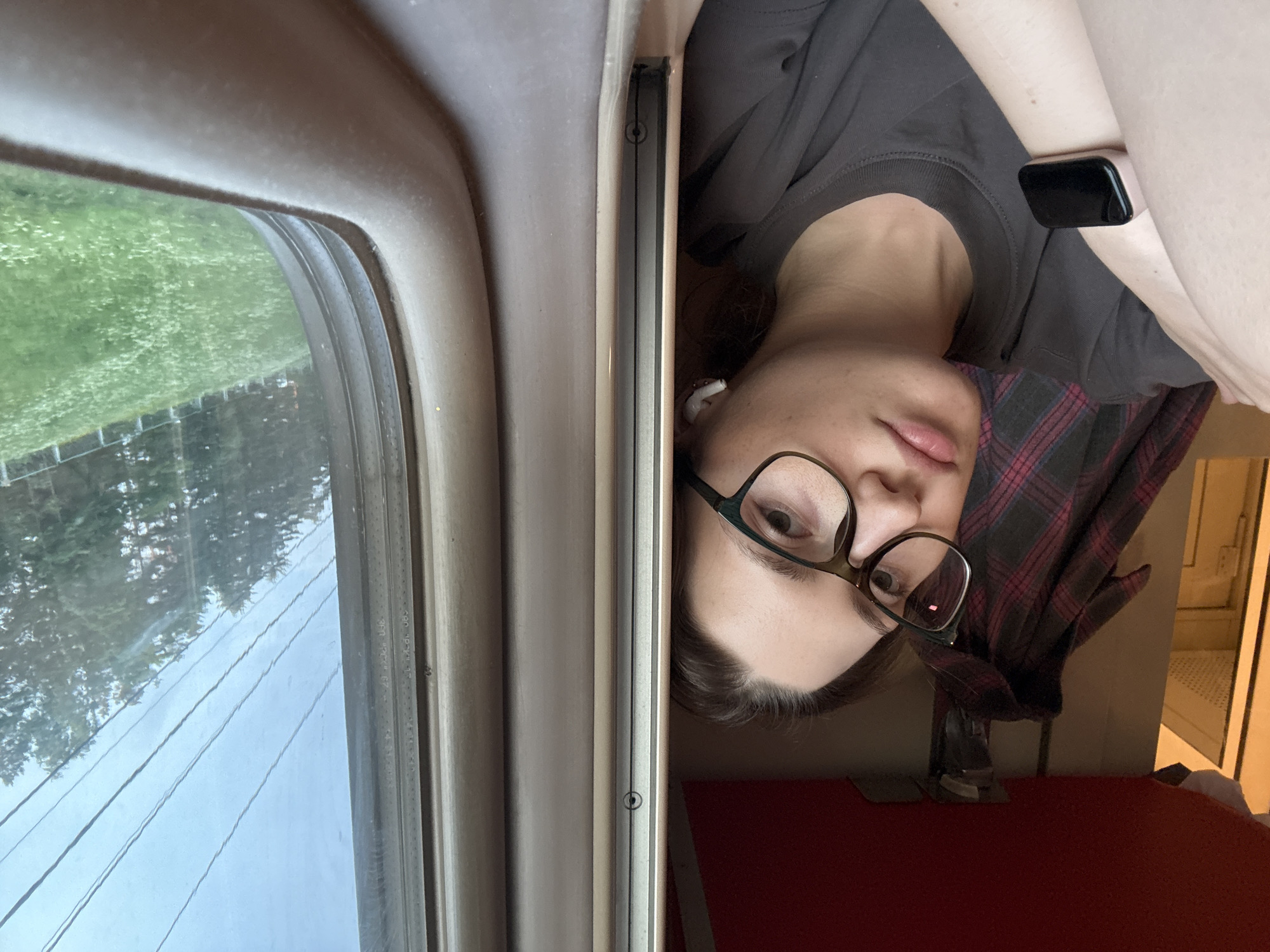 A young woman with glasses and brown hair, wearing a grey t-shirt and a smartwatch on her left wrist, is seen from the chest up, looking towards the camera. She is in a train compartment, with a red ceiling visible above and a plaid garment hanging on the wall behind her. To her right, a window shows a green, grassy landscape with trees and power lines outside, suggesting a rural or suburban setting. The scene's context is Povarovo, Russia, indicating the location of the train journey.