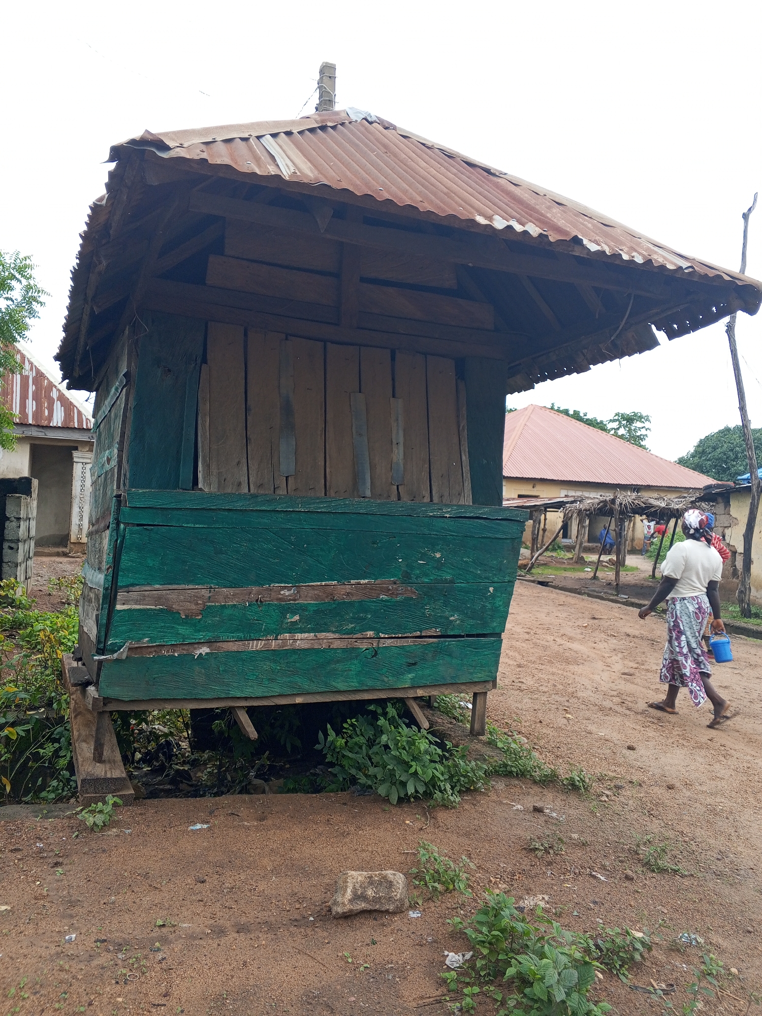 In the city of Nyita, Nigeria, a weathered, green wooden kiosk stands in the foreground, its corrugated metal roof showing signs of rust. The kiosk appears elevated on wooden supports, with dense green foliage growing underneath. To the right of the kiosk, a woman in a patterned skirt and white top walks away from the camera, carrying a small blue bucket in her right hand. The ground is a reddish-brown dirt path, scattered with small pebbles and patches of sparse vegetation. In the middle ground, several other buildings with corrugated metal roofs are visible, one with a more substantial structure and a covered porch supported by wooden posts. A few other individuals are loosely gathered near this building. The sky is overcast, suggesting a cloudy day.