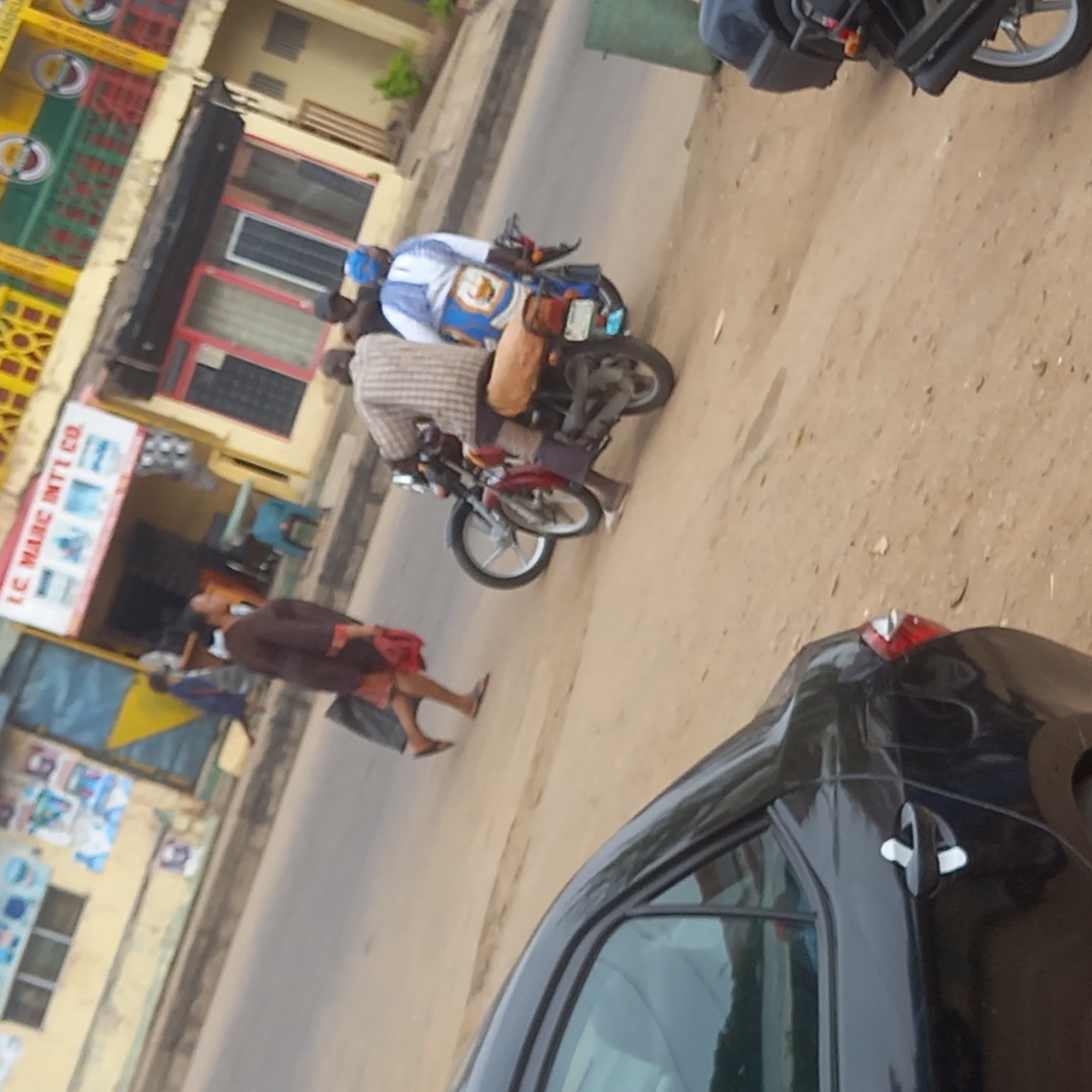 The scene is in Dadi Riba, Nigeria, showing a street setting during daylight. A black car is parked on the right side of the image. A woman in a brown dress and carrying a black bag is walking on the sidewalk. Two people are riding motorcycles in the street. A building with a sign that reads "I.C. MARC INTI CO." is visible on the left. The building facade is adorned with yellow, green, and red crate structures.
