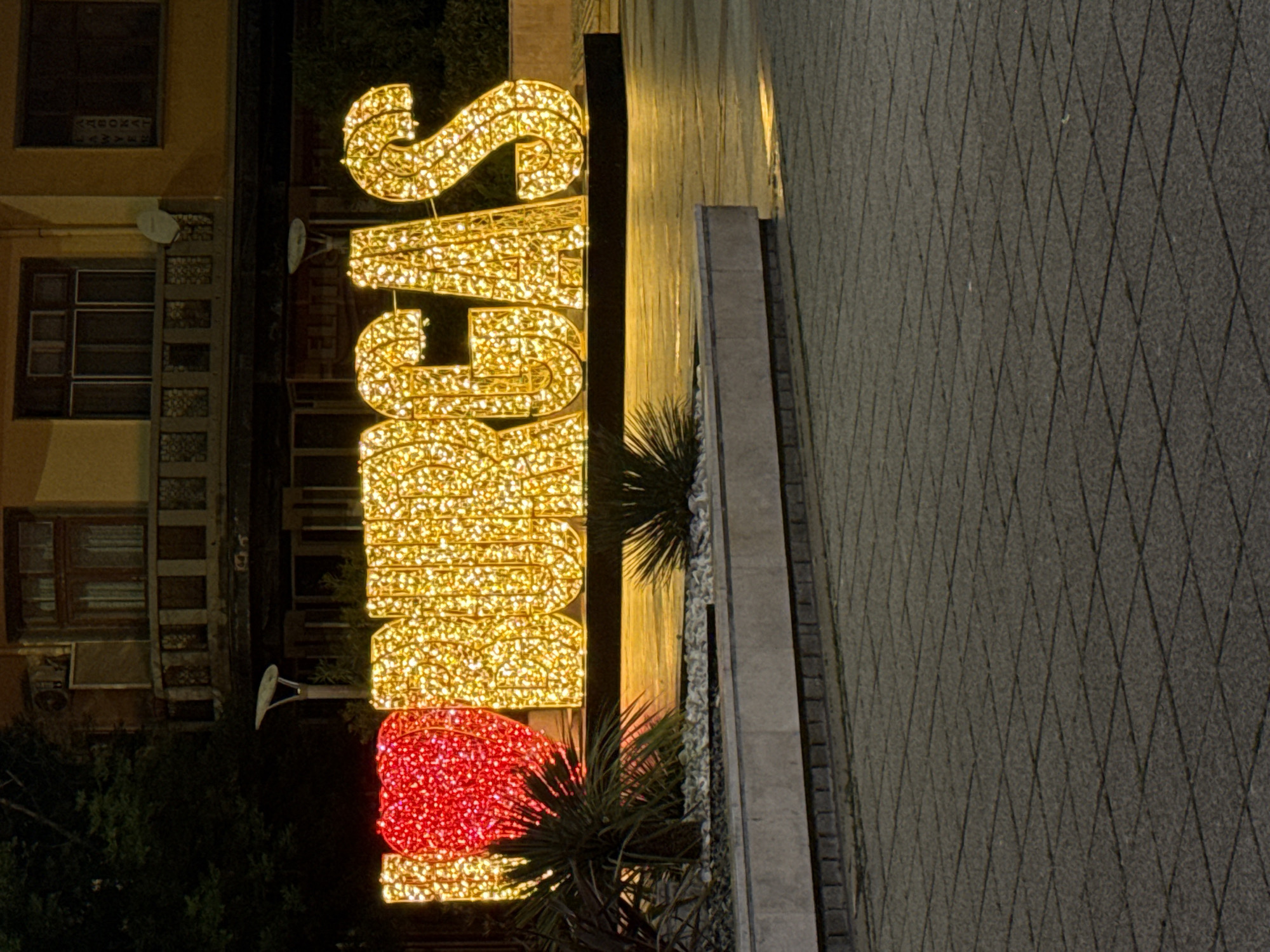 A large, illuminated sign that reads "I LOVE BURGAS" is situated in an outdoor urban setting. The sign consists of individual letters, with the "I" represented by a large, red heart shape and the letters "LOVE BURGAS" formed by a dense arrangement of warm, yellow-orange lights. The sign is elevated and appears to be a permanent installation. In front of the sign, there are low-lying green plants and a stone border. The ground in the foreground is paved with square tiles, and in the background, a building with balconies and windows is visible. The lighting suggests it is either dusk or nighttime, with artificial lights prominently featured. There are no people or animals in the image.