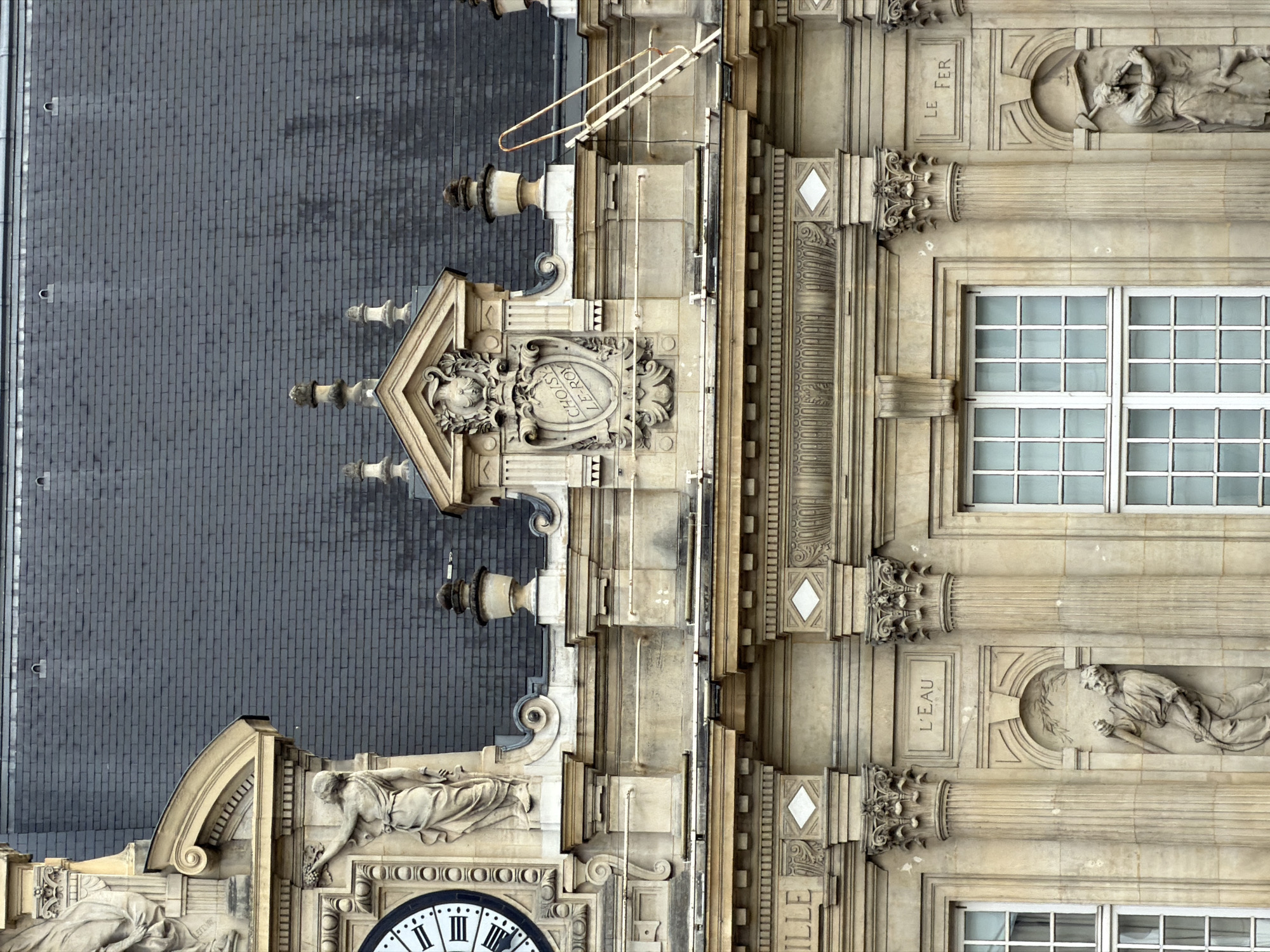This media file captures an upward-facing, eye-level, full shot of the facade of the Hotel de Ville in Ivry-sur-Seine, France. The architectural style is classical, with detailed carvings and sculptures adorning the stone building. The facade is divided vertically by fluted columns and horizontally by decorative bands.
On the left side of the image, a dark grey, textured roof made of what appears to be slate tiles occupies a significant portion. Scattered across the roof are various architectural elements, including dormer windows and finials. To the right of the roof, a section of the building's facade is visible, featuring ornate carvings. One prominent element is a shield-like crest with a face and the inscription "CHOISIR LE ROY." Above this crest, a set of metal railings suggests a maintenance platform or decorative feature.
The central and right portions of the image showcase the main facade, characterized by a lighter, beige-colored stone. Large, multi-paned windows with white frames are visible on this section. Numerous sculptures are integrated into the facade, depicting figures in various poses. One sculpture shows a reclining figure holding a staff and a wreath, with the inscription "L'EAU" nearby. Another sculpture, above a window, shows a figure with a tool, and the inscription "LE FER" is also visible. Below these, a large clock face with Roman numerals is partially visible at the bottom of the frame. The time on the clock appears to be approximately 2:20.
The lighting suggests it is daytime, with soft shadows indicating an overcast or diffused light condition. There are no clear indications of weather or specific events taking place, creating a static, architectural study. The overall impression is one of historical grandeur and detailed craftsmanship.